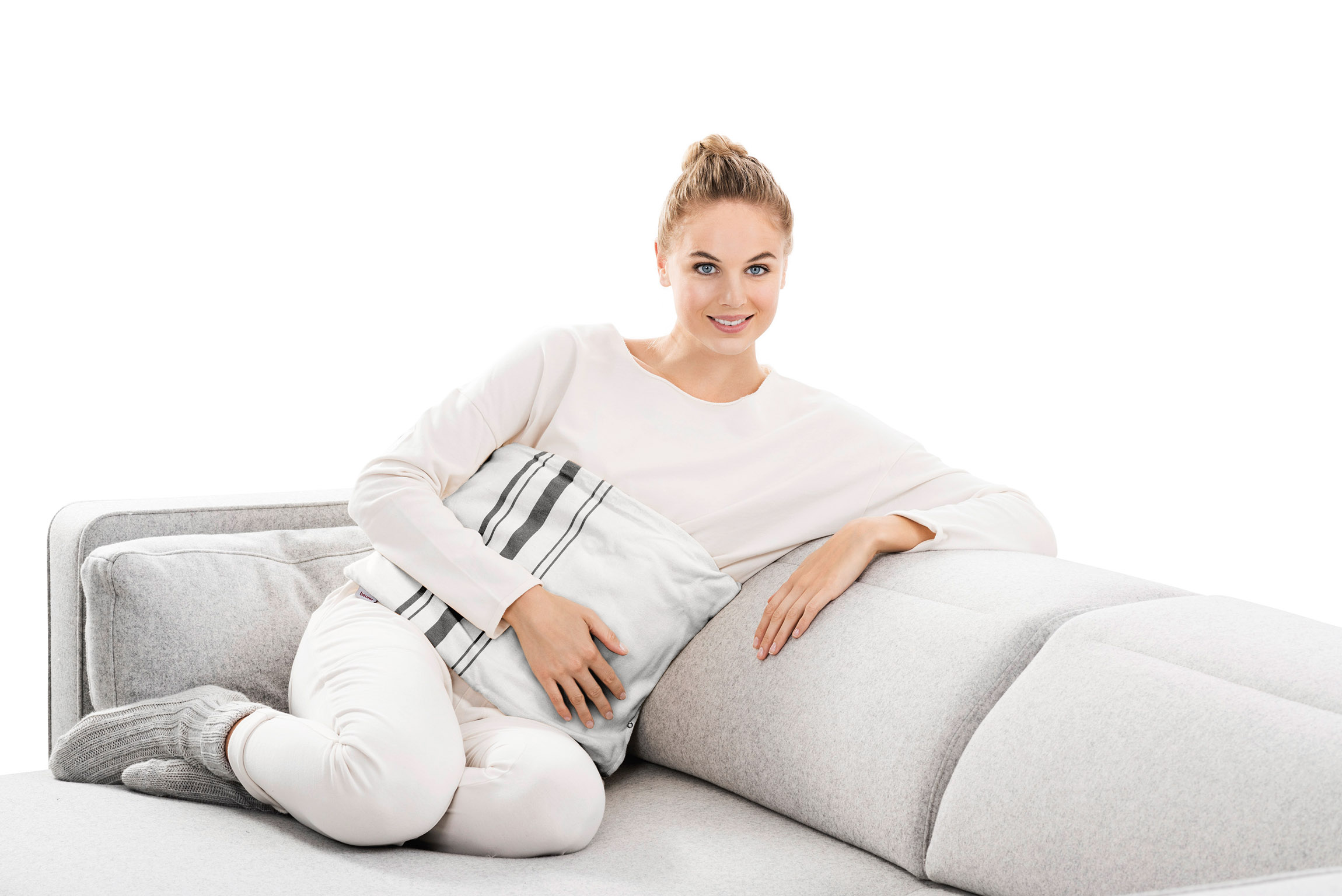 Eine Frau sitzt auf einem grauen Sofa, hält ein Kissen und lächelt in die Kamera.