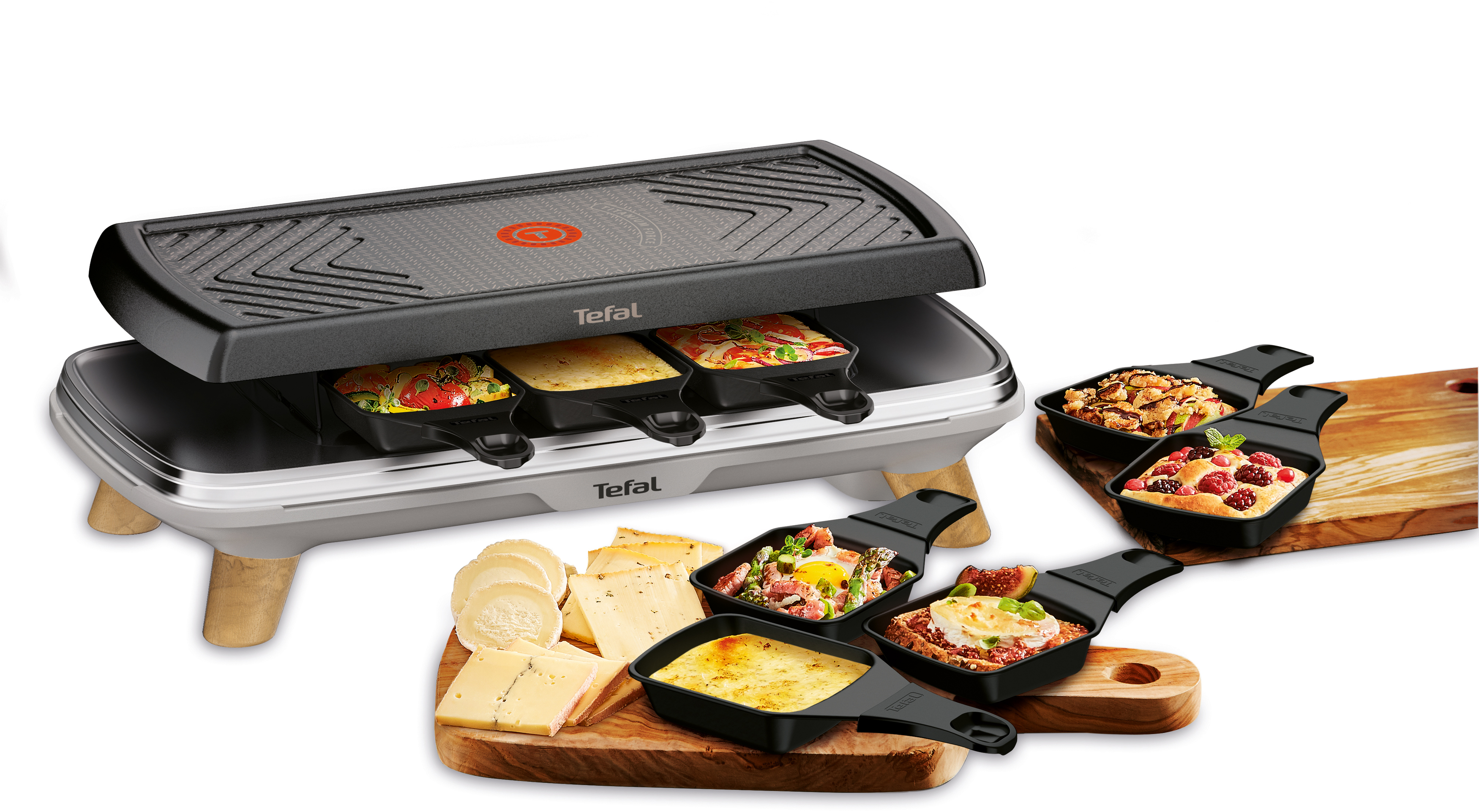 Raclette-Grill mit Essen. Enthält Käse, Gemüse und kleine Pfannen mit Griffen. Weißer Hintergrund.