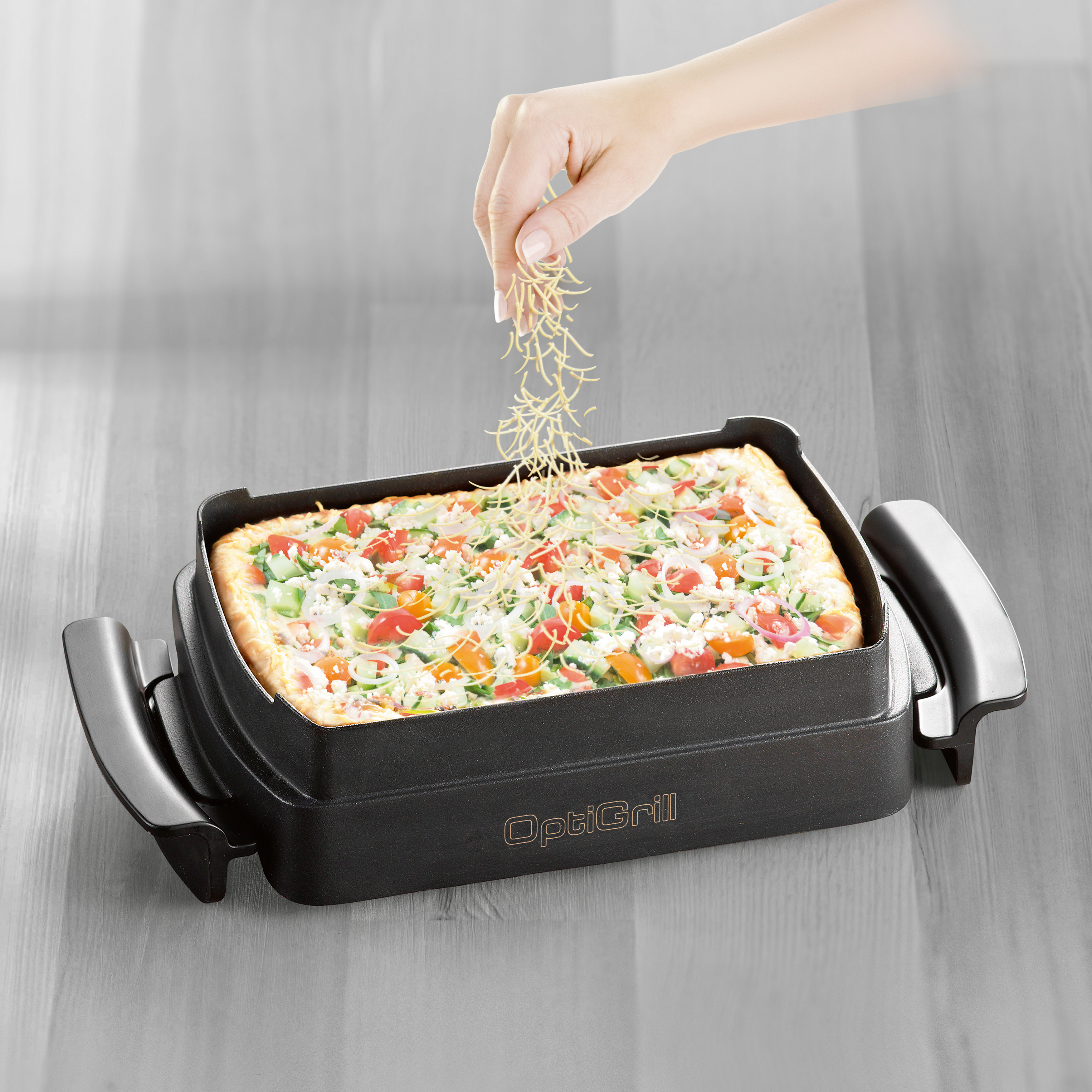 Pizza in einem schwarzen OptiGrill, Hand streut Käse oben drauf, auf hellem Holz.