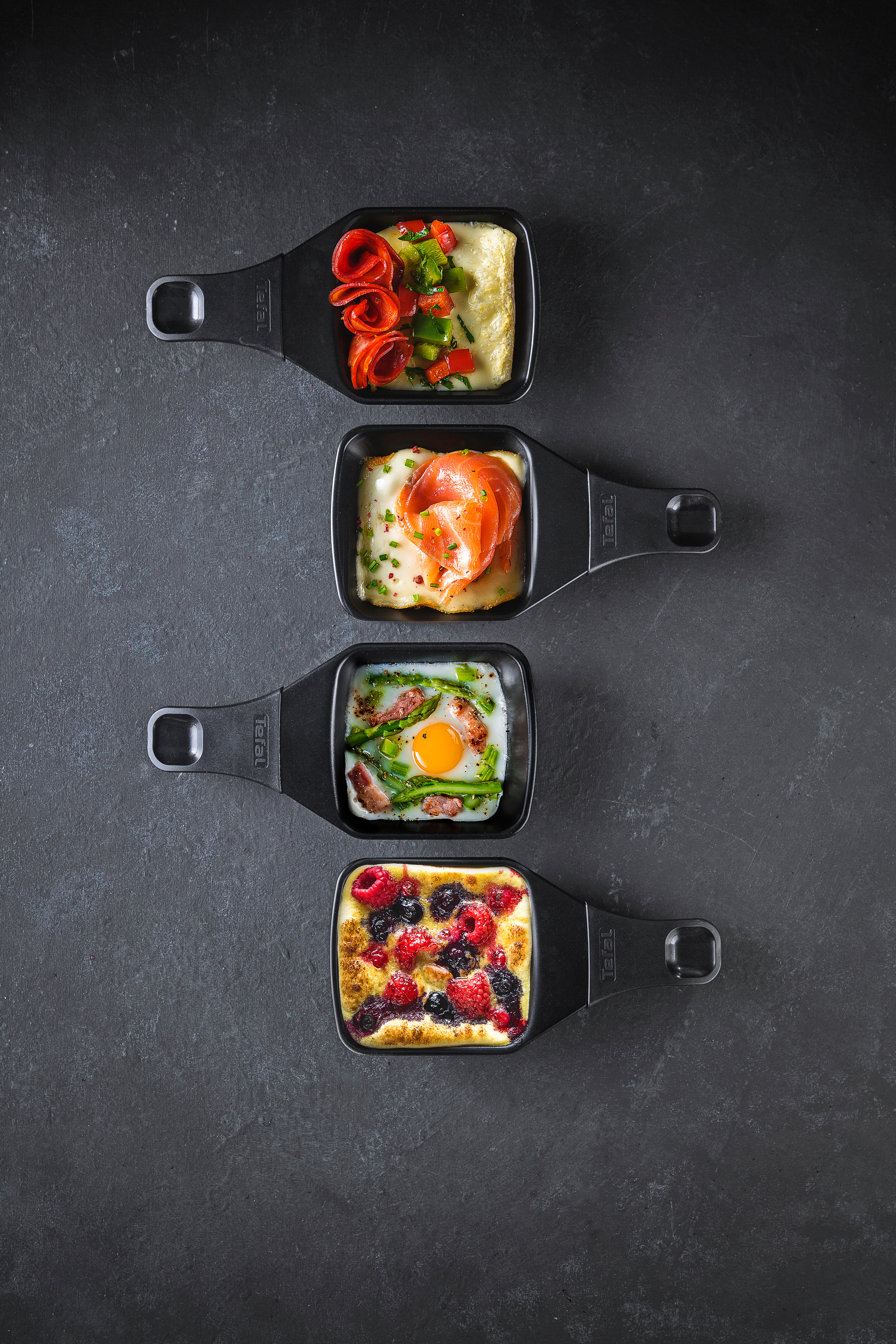 Vier Mini-Pfannen mit verschiedenen gekochten Speisen. Sie stehen auf einer dunklen Oberfläche, mit Lachs, Ei und Beeren.