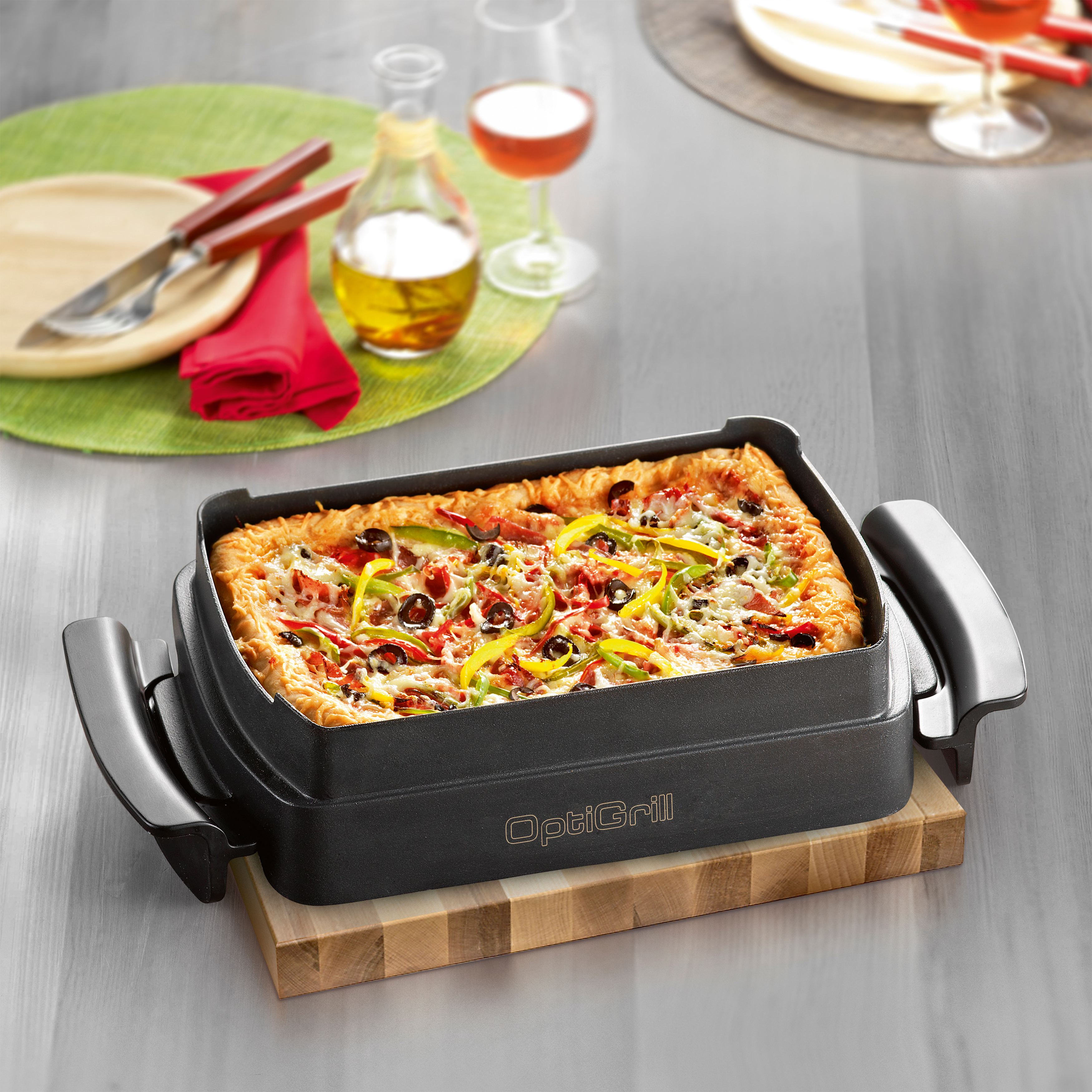 Eine rechteckige Pizza in einer dunklen Pfanne auf einem Holzbrett. Weingläser und eine Ölflasche sind im Hintergrund.