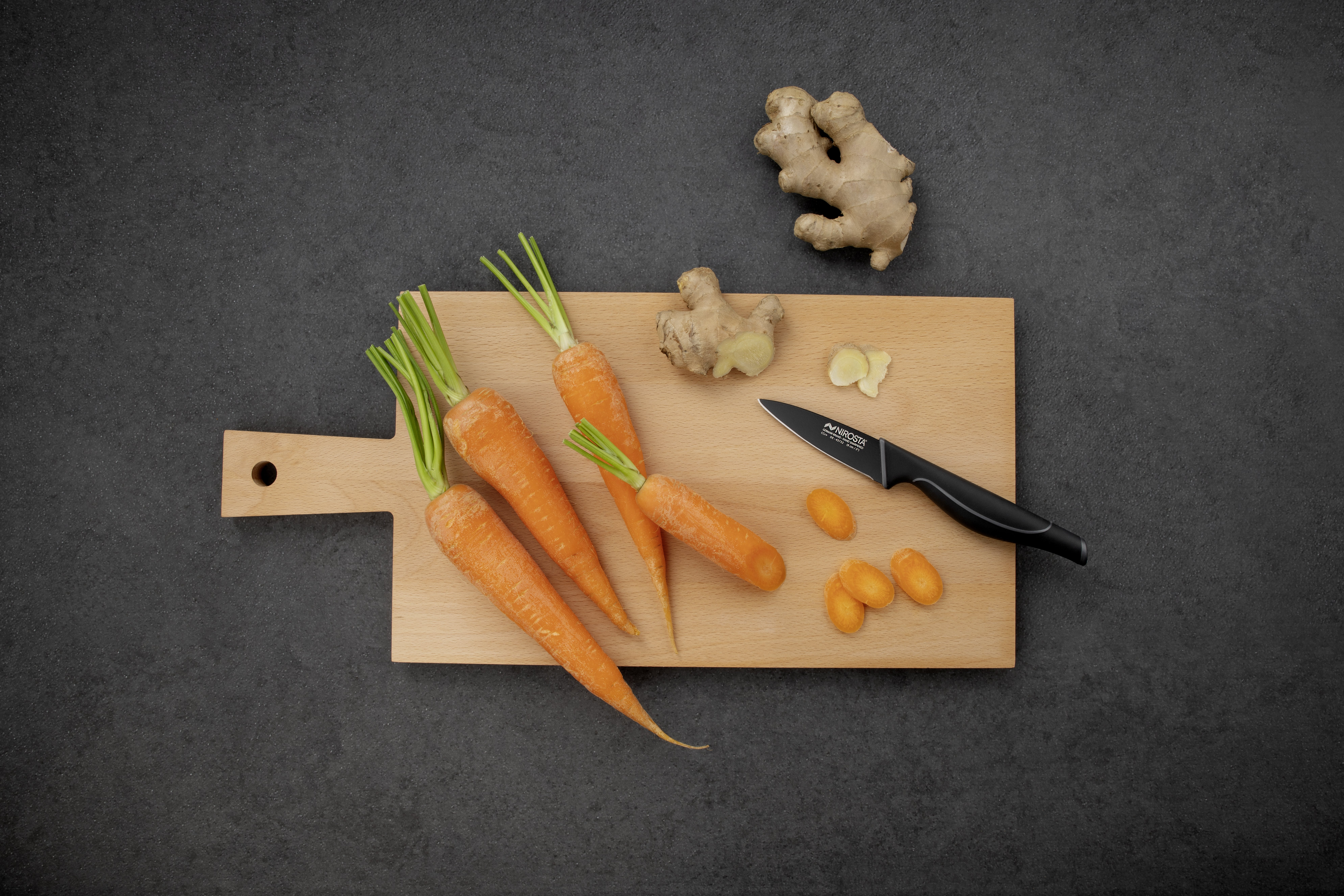 Karotten, Ingwer, ein schwarzes Messer und Kumquats auf einem Holzschneidebrett. Dunkelgrauer Hintergrund.