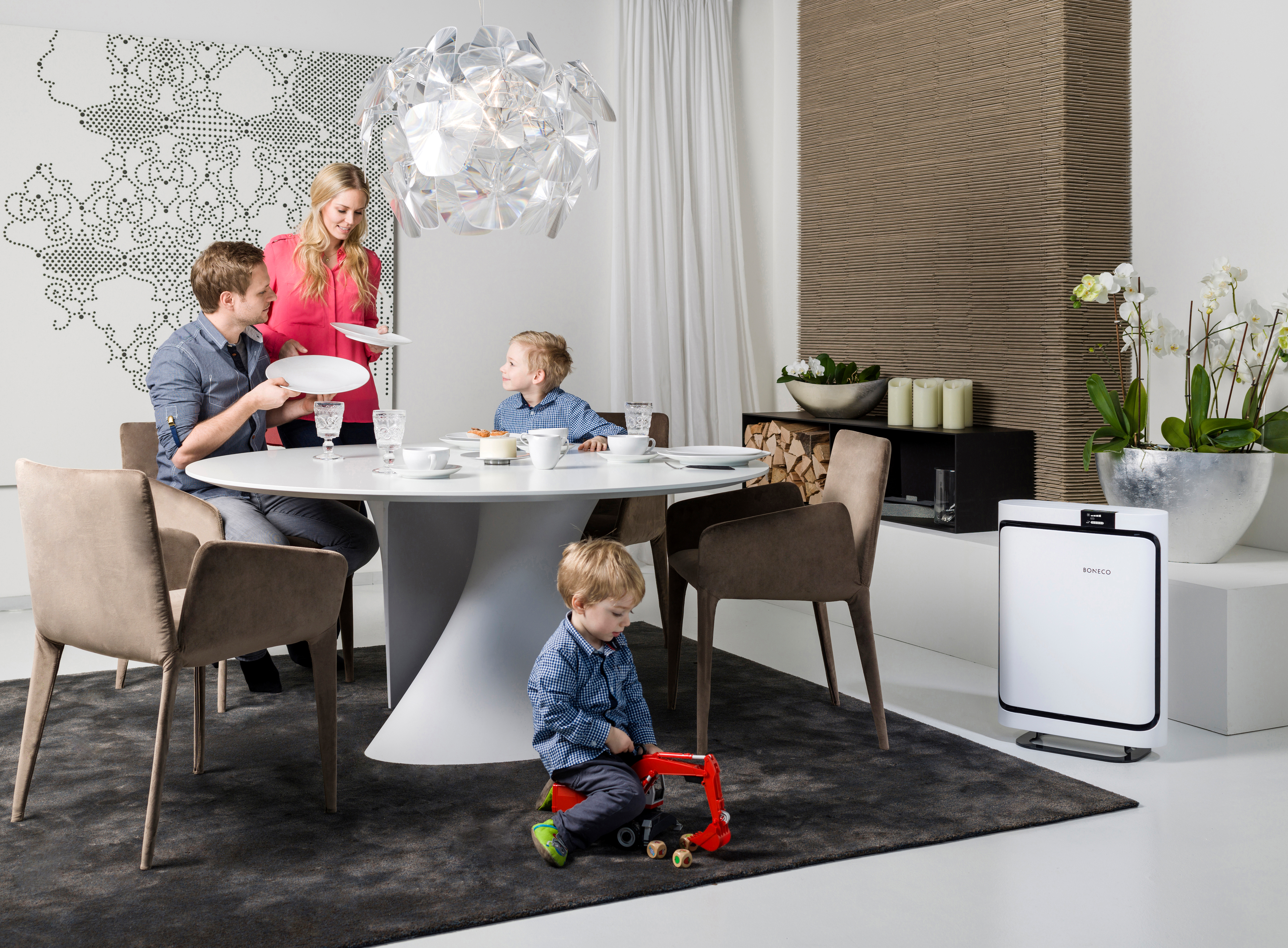 Familie in einem Esszimmer. Eltern decken den Tisch. Kleinkind spielt mit einem Spielzeug. Weißer Luftreiniger.