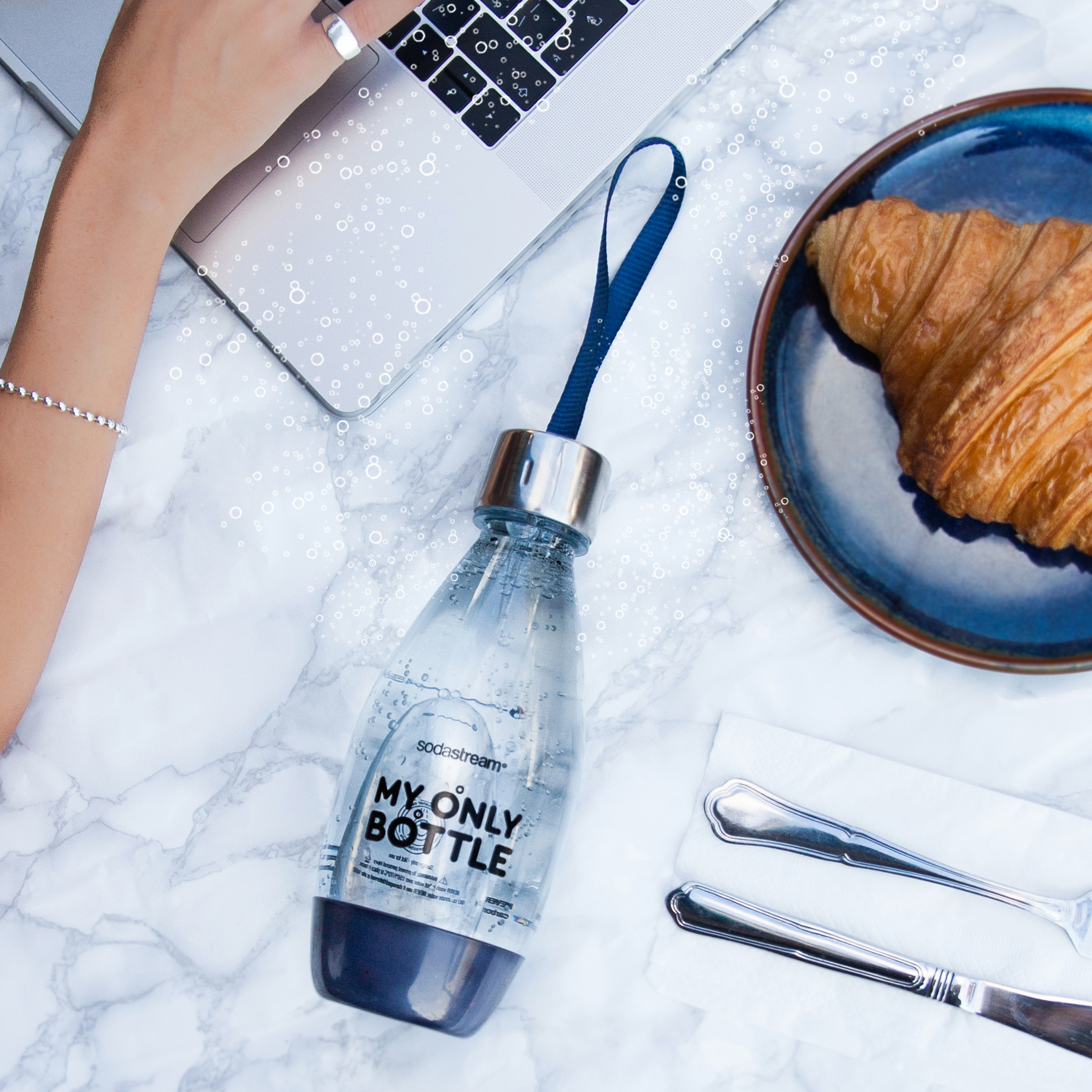 Une bouteille sur une surface en marbre avec un ordinateur portable, un croissant et des couverts. Vue de dessus.