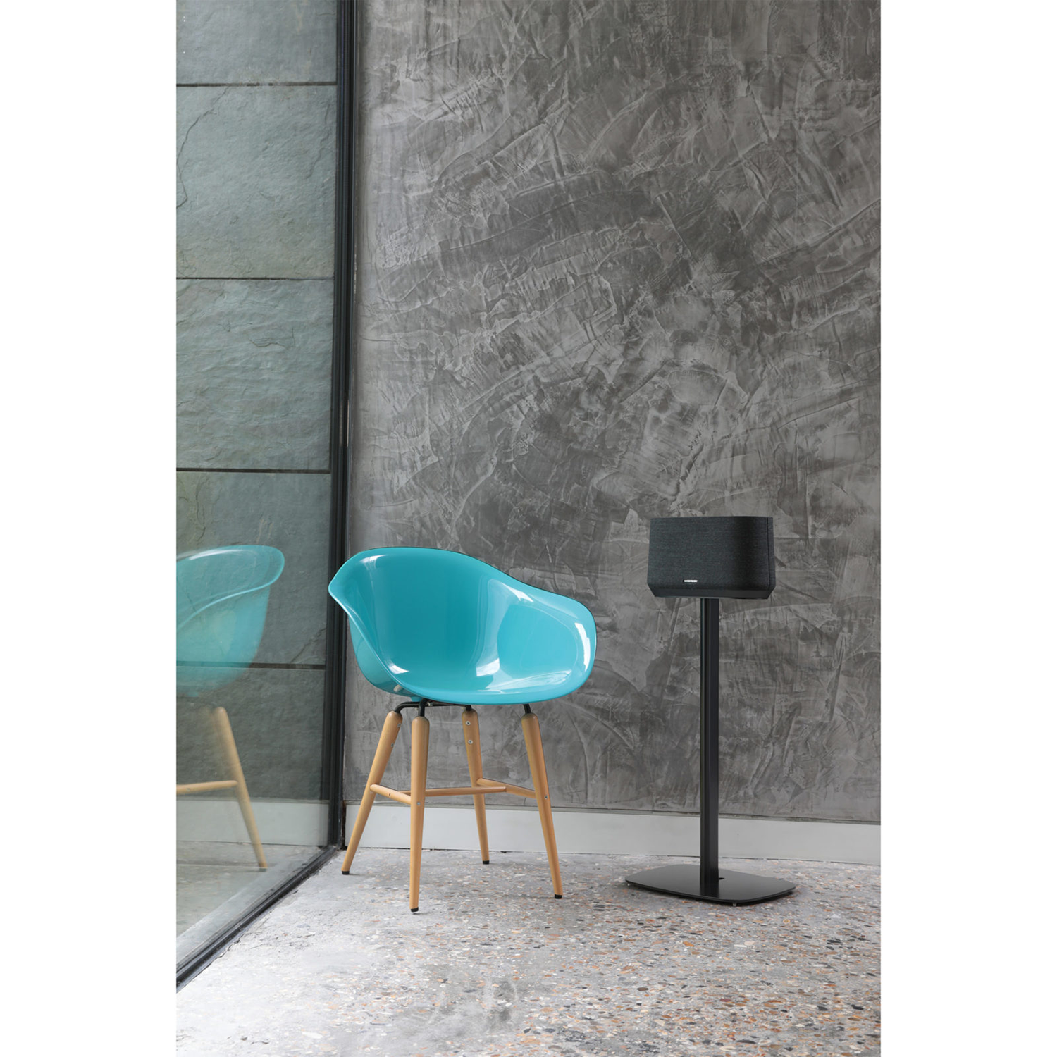 Een blauwe stoel, een speaker op een standaard en een grijze getextureerde muur. Achtergrond is een raam.