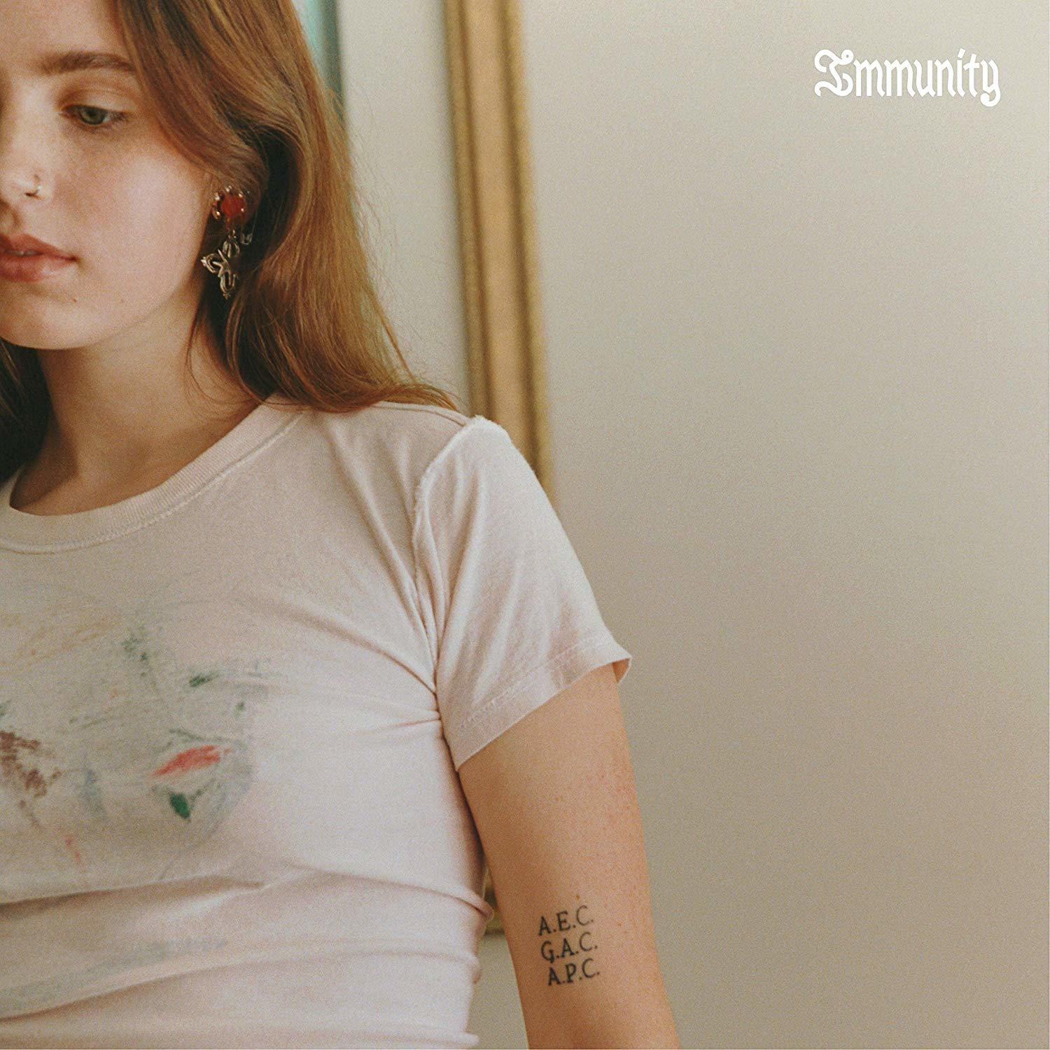 Eine junge Frau mit roten Haaren und Nasenring, trägt ein weißes Hemd. Ein Tattoo ist auf ihrem Arm sichtbar.