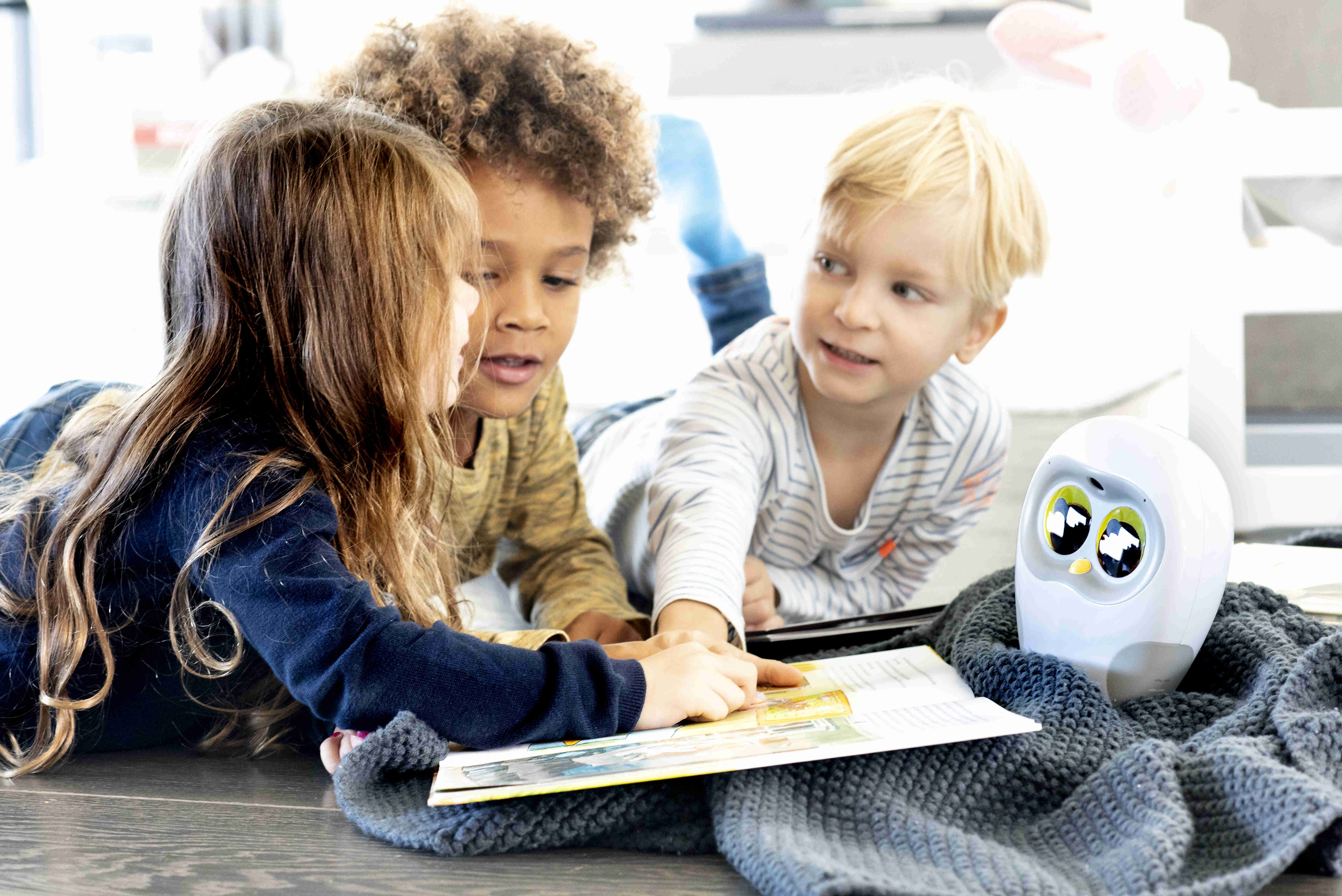 Drei Kinder lesen gemeinsam ein Buch auf dem Boden, mit einem weißen, eulenartigen Roboter.