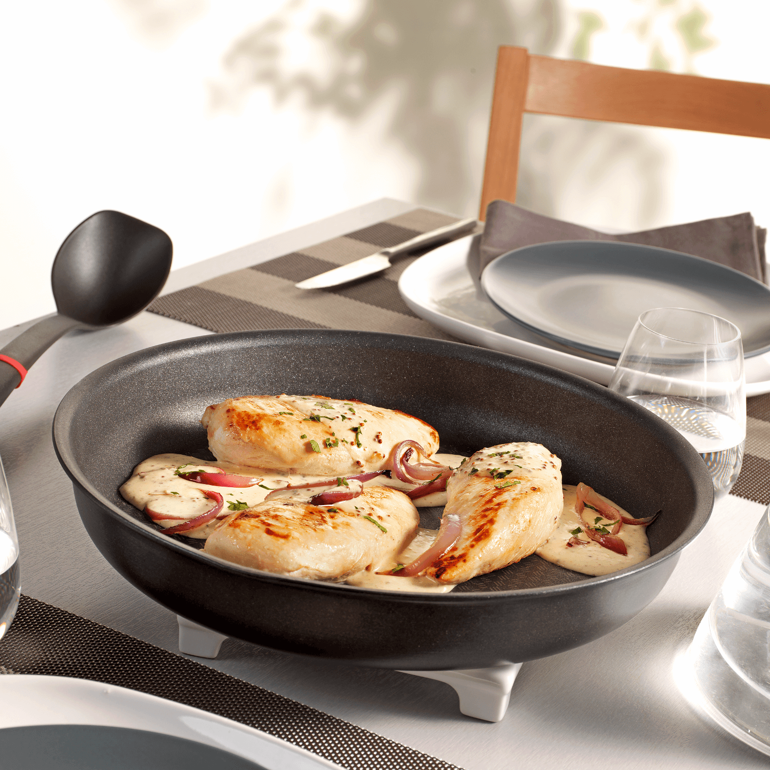 Kipfilets in een pan met saus en rode ui, geserveerd op een tafel met borden en glazen.