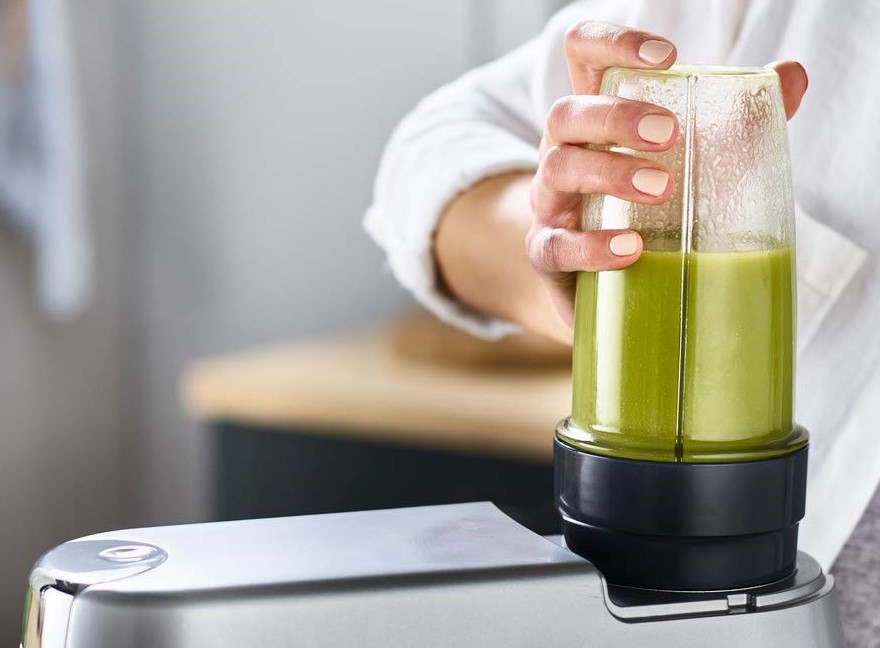 Hand hält Blenderbehälter mit grüner Flüssigkeit, silberner Basis.
