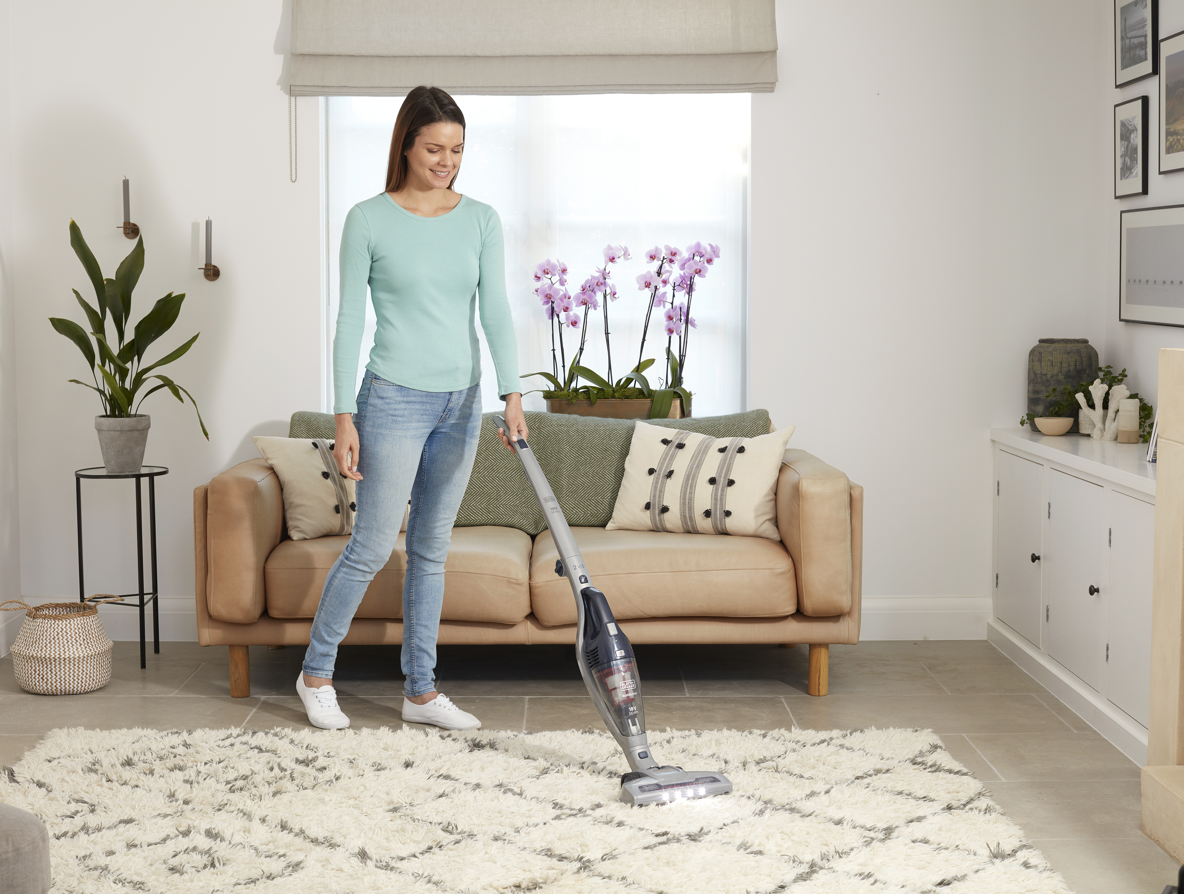 Frau, die einen weißen Shag-Teppich mit einem kabellosen Staubsauger saugt. Innenraum mit Pflanzen und Sofa.