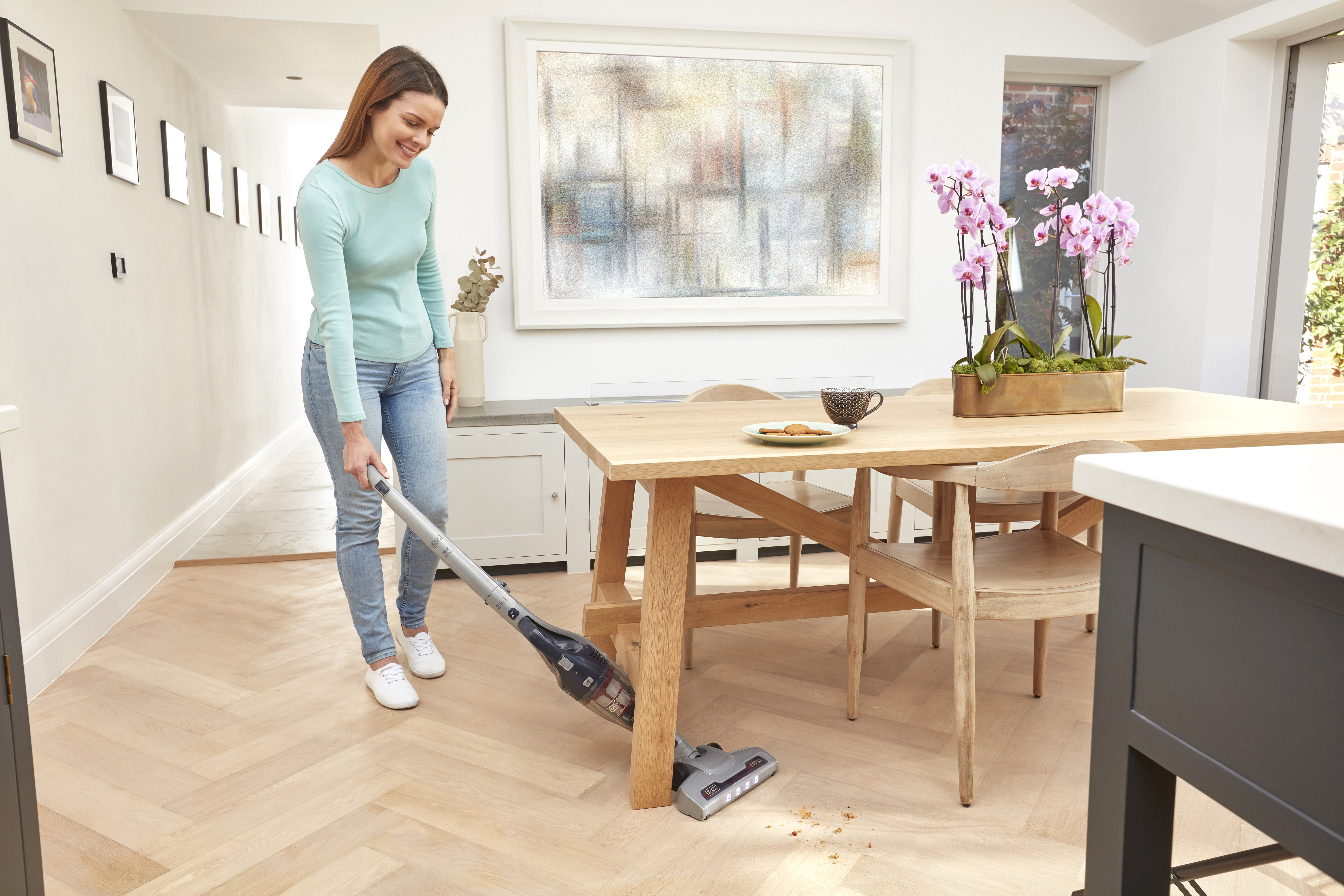 Frau saugt Holzboden im Essbereich. Tisch mit Essen und Blumen hinter ihr.