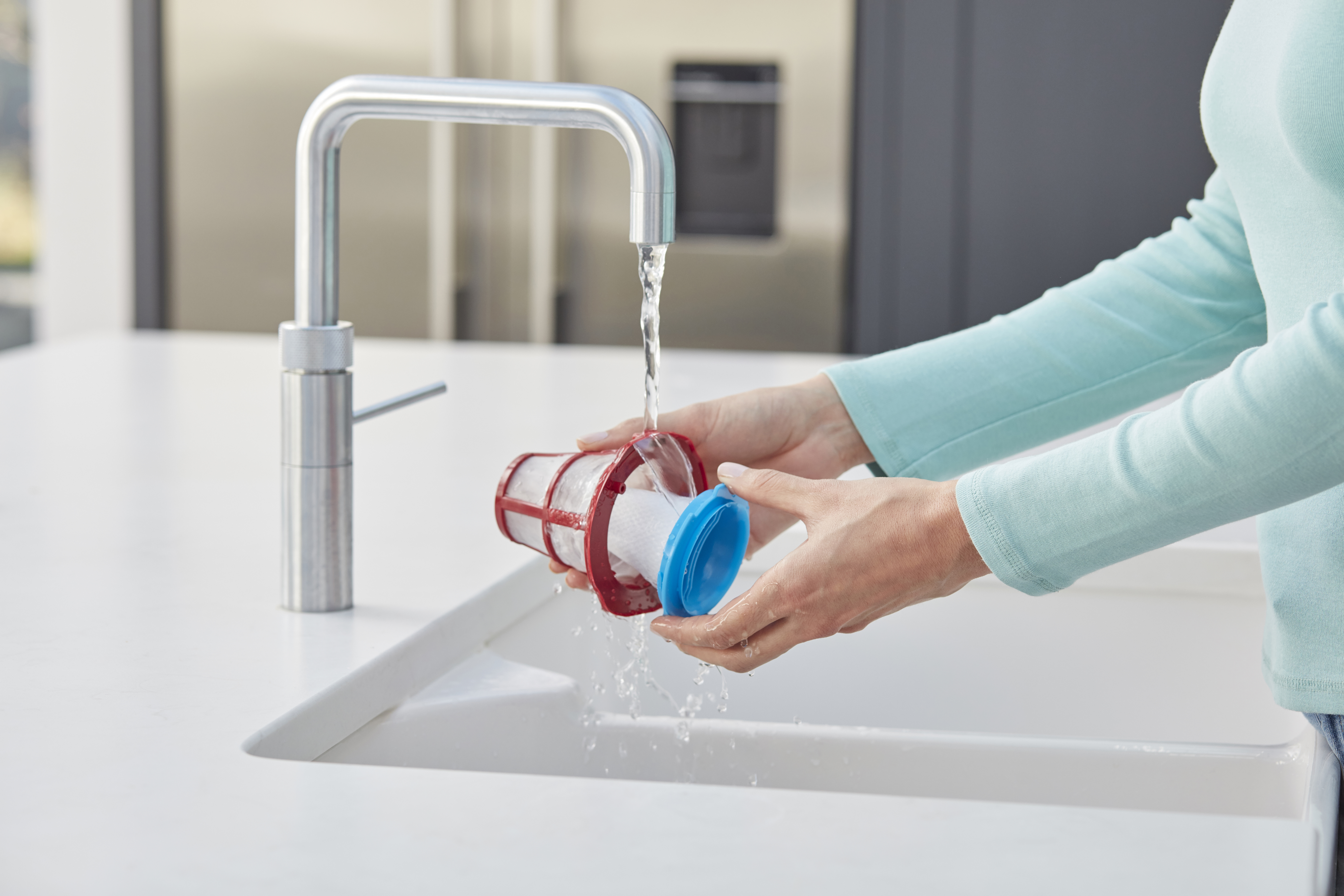 Eine Frau wäscht den Filter eines Staubsaugers unter einem Wasserhahn. Der Filter ist rot und blau.