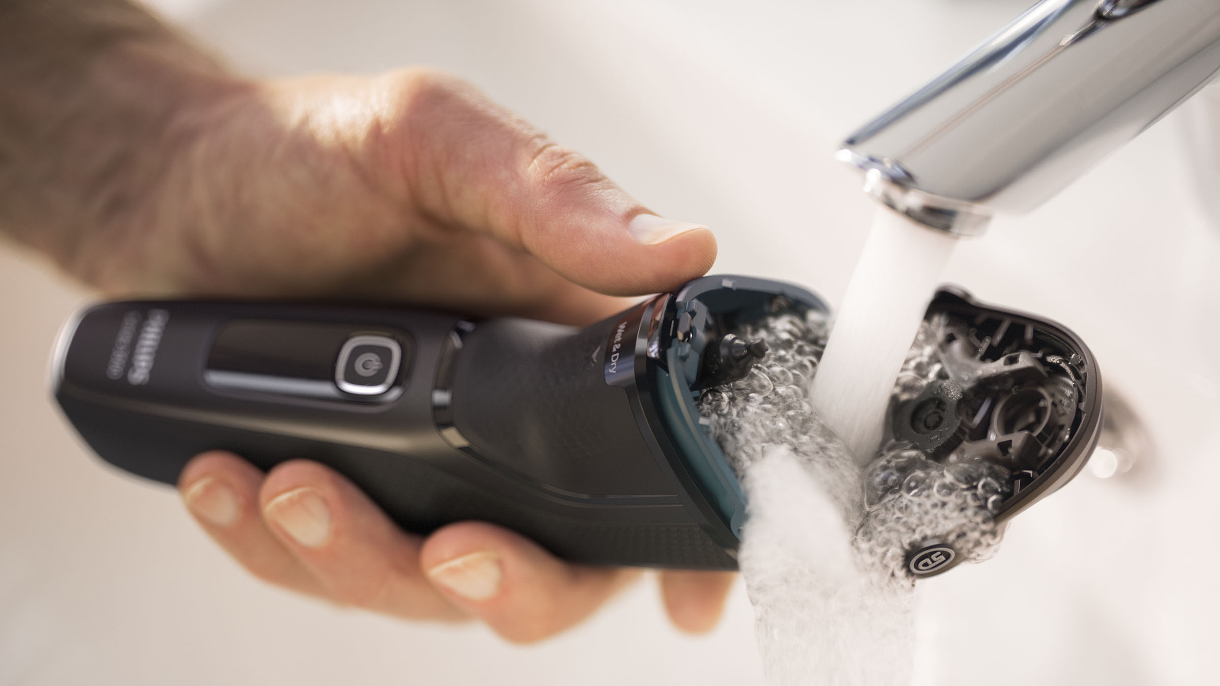 Eine Person spült einen schwarzen Elektrorasierer unter fließendem Wasser ab. Der Rasierer ist zum Reinigen geöffnet.