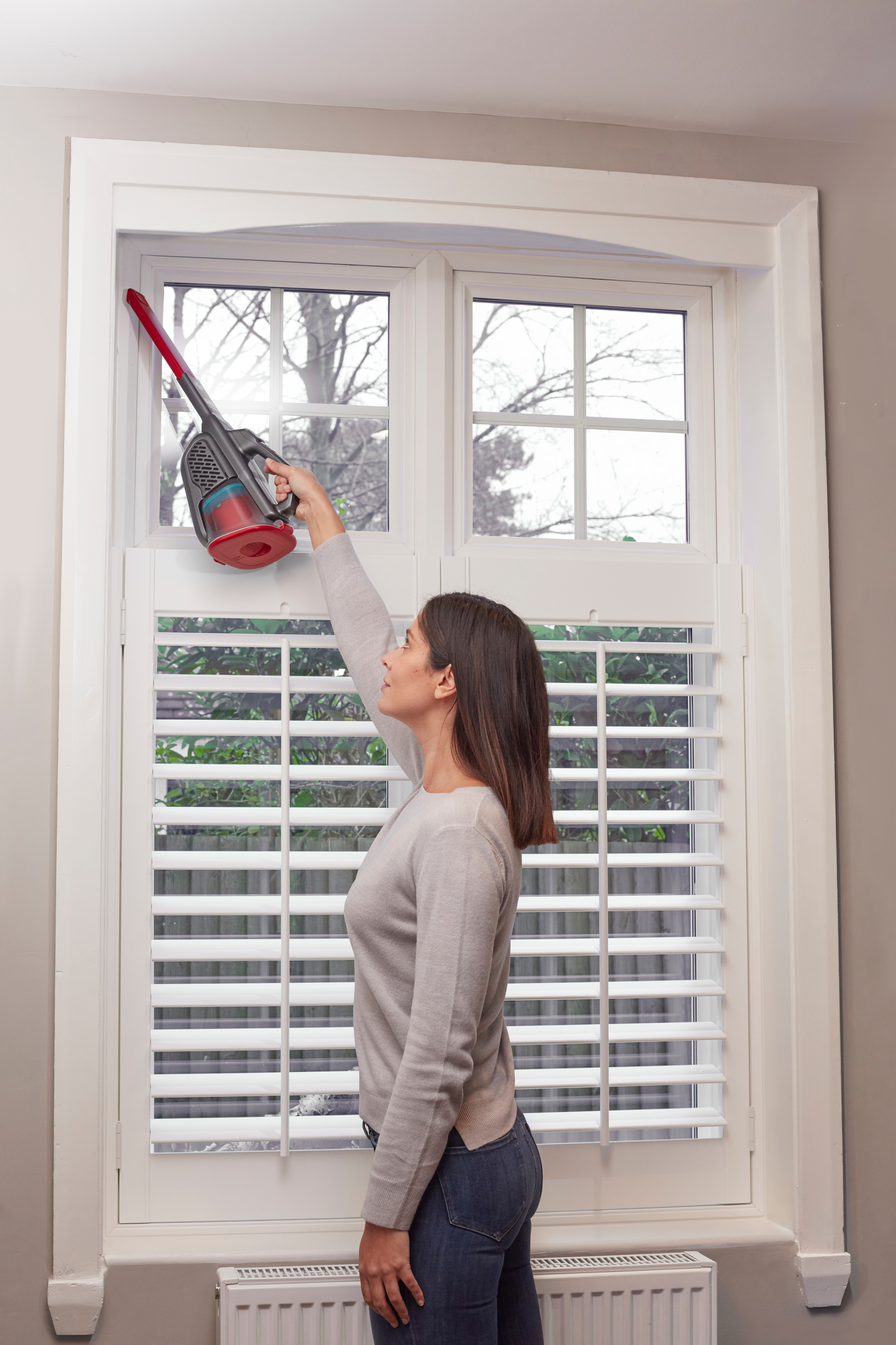 Frau, die Fensterläden saugt. Der Staubsauger ist rot und grau. Das Fenster ist weiß, mit Fensterläden.