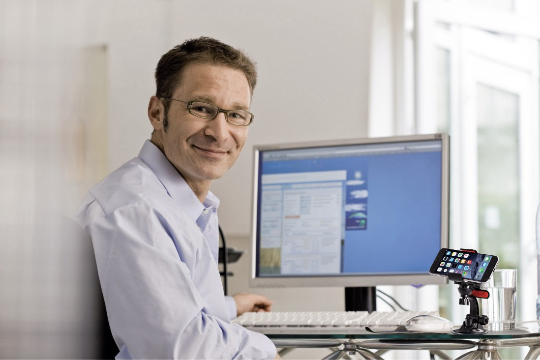 Homme en lunettes à l'ordinateur sourit, téléphone sur support. Fond : bureau, fenêtre.