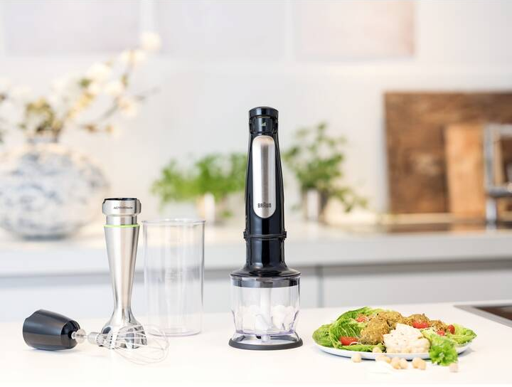 Mixer mit Aufsätzen, Glas und Essen auf einer weißen Arbeitsplatte. Grüne Pflanzen im Hintergrund.