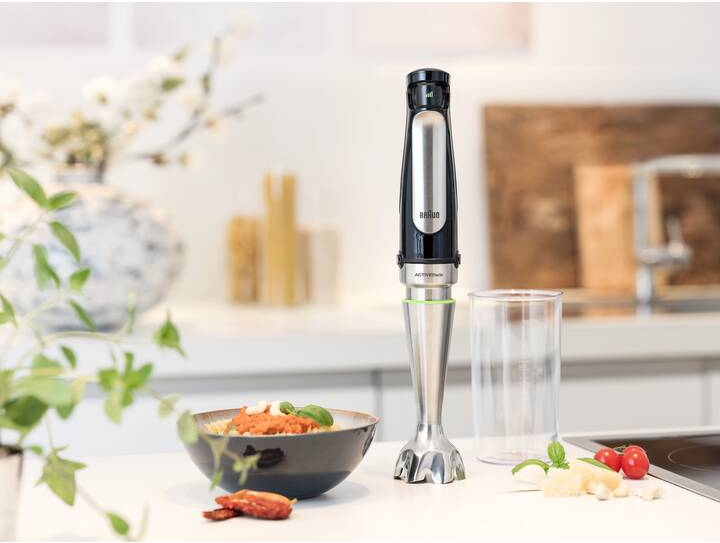 Stabmixer mit Essen in einer Schüssel und Zutaten auf einer weißen Arbeitsplatte, helle Beleuchtung.