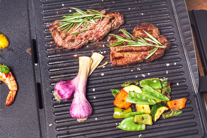 Gegrillte Steaks mit Rosmarin, Knoblauchzehen und gegrilltem Gemüse auf einem schwarzen Grill.