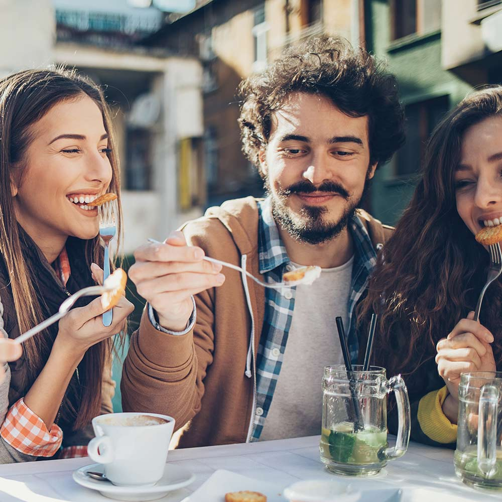 Drei Personen am Tisch, essen Essen mit Gabeln. Getränke und eine weiße Kaffeetasse sind vorhanden.