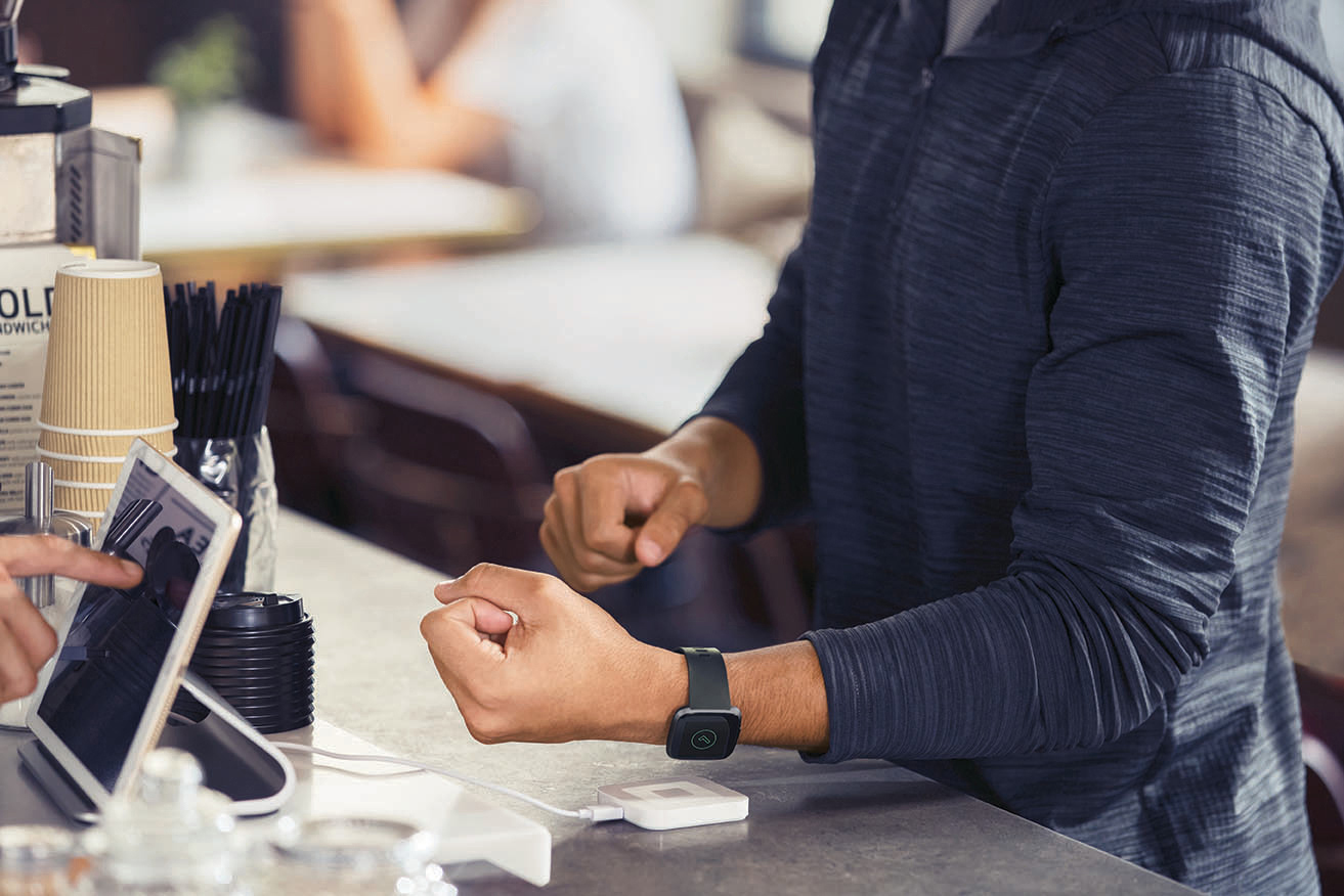 Person zahlt mit Smartwatch am Tresen, Kartenlesegerät vorhanden, drinnen.