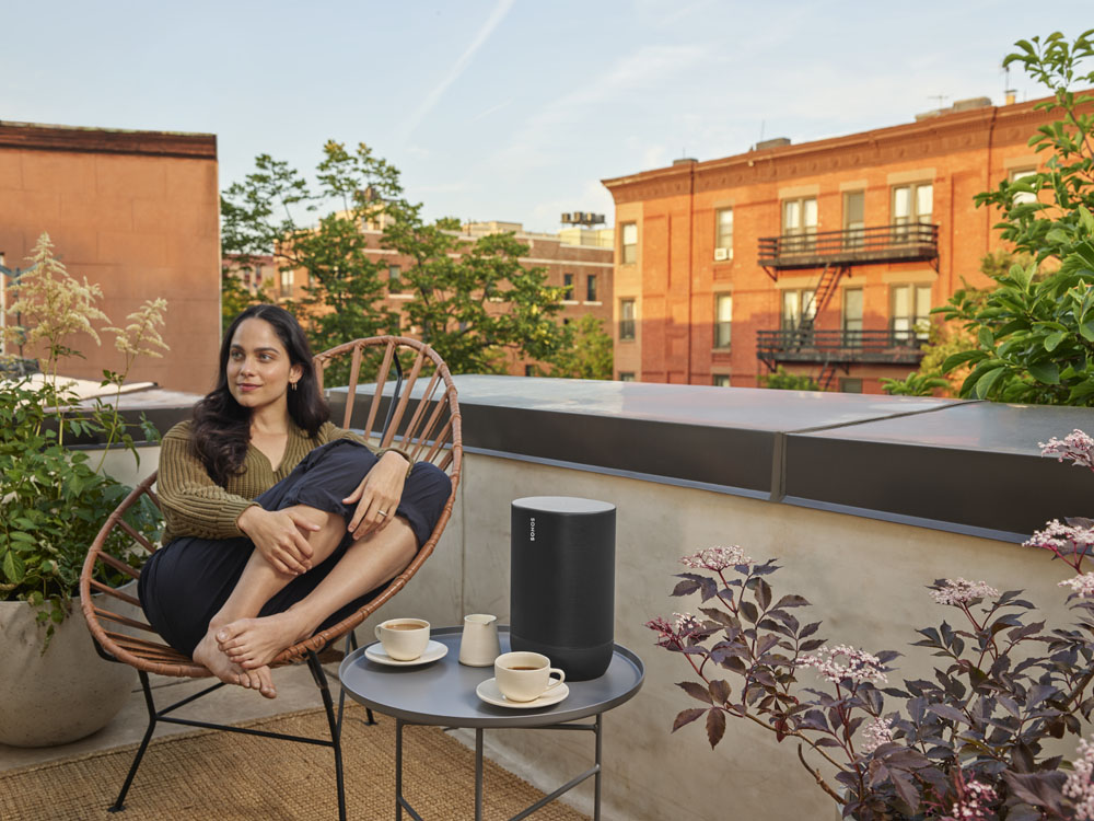 Een vrouw zit op een balkon. Ze zit in een rieten stoel. Een luidspreker staat op een tafel. Gebouwen op de achtergrond.