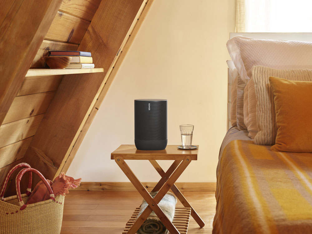 Een zwarte luidspreker op een houten tafel naast een glas water. Een bed en boeken op de achtergrond.