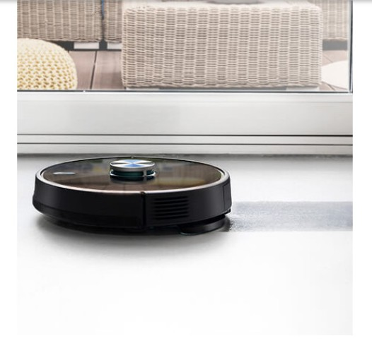Aspiradora robot negra limpiando piso blanco, cerca de puerta de vidrio.