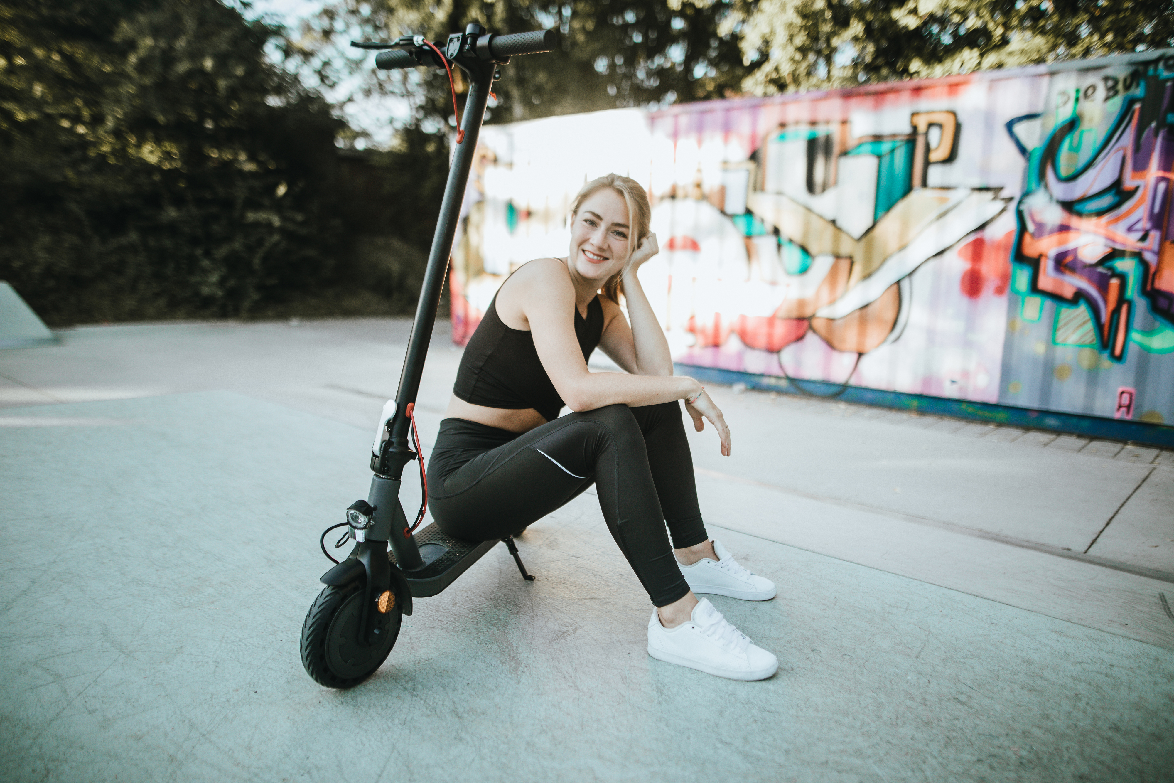 Blonde Frau sitzt auf einem schwarzen Elektroroller und lächelt. Graffiti-Kunst im Hintergrund.