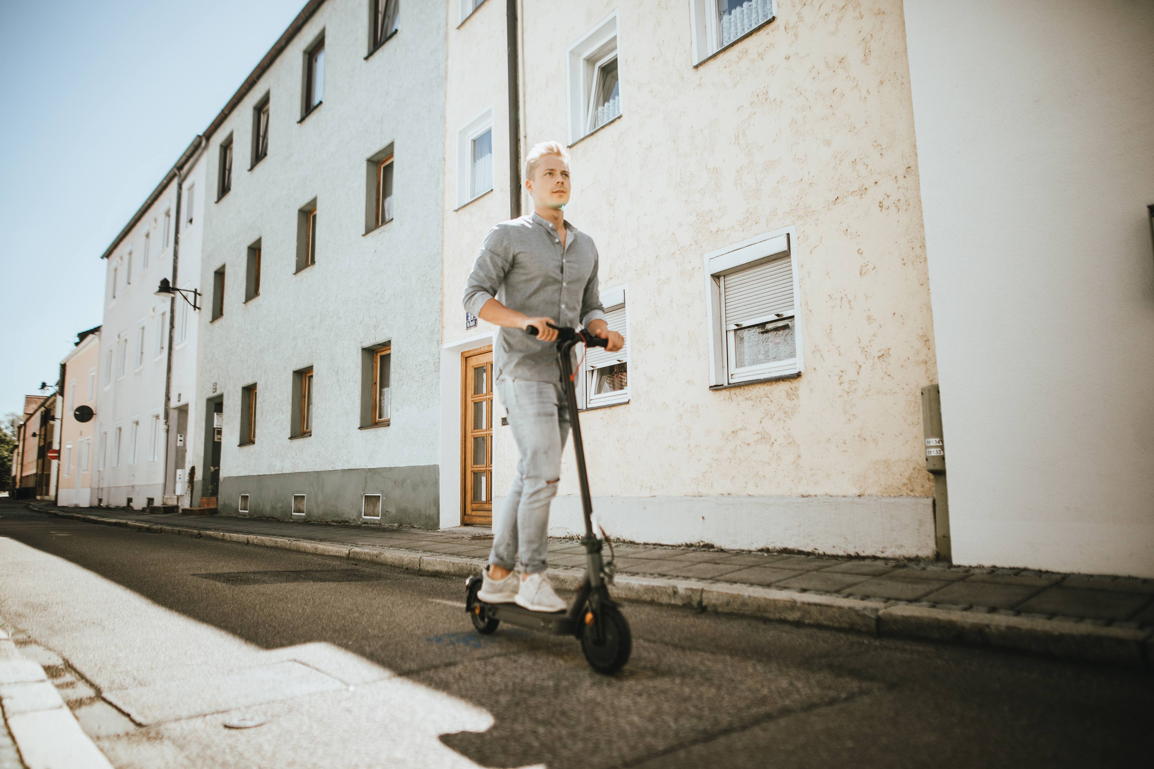 Ein Mann fährt mit einem Elektroroller eine Straße entlang, Gebäude beidseitig.