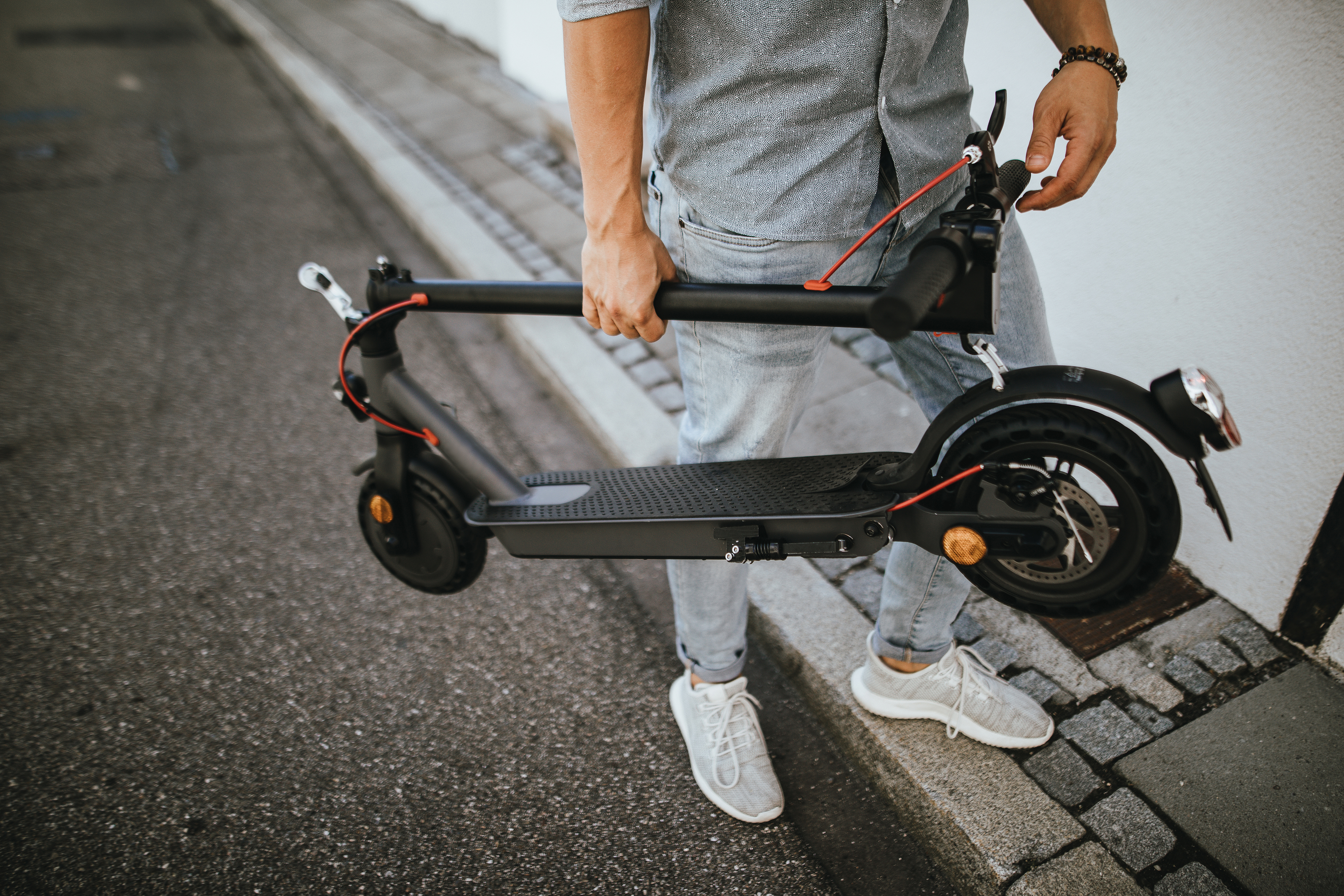 Person hält zusammengeklappten schwarzen Roller auf der Straße, graue Hose, weiße Schuhe.