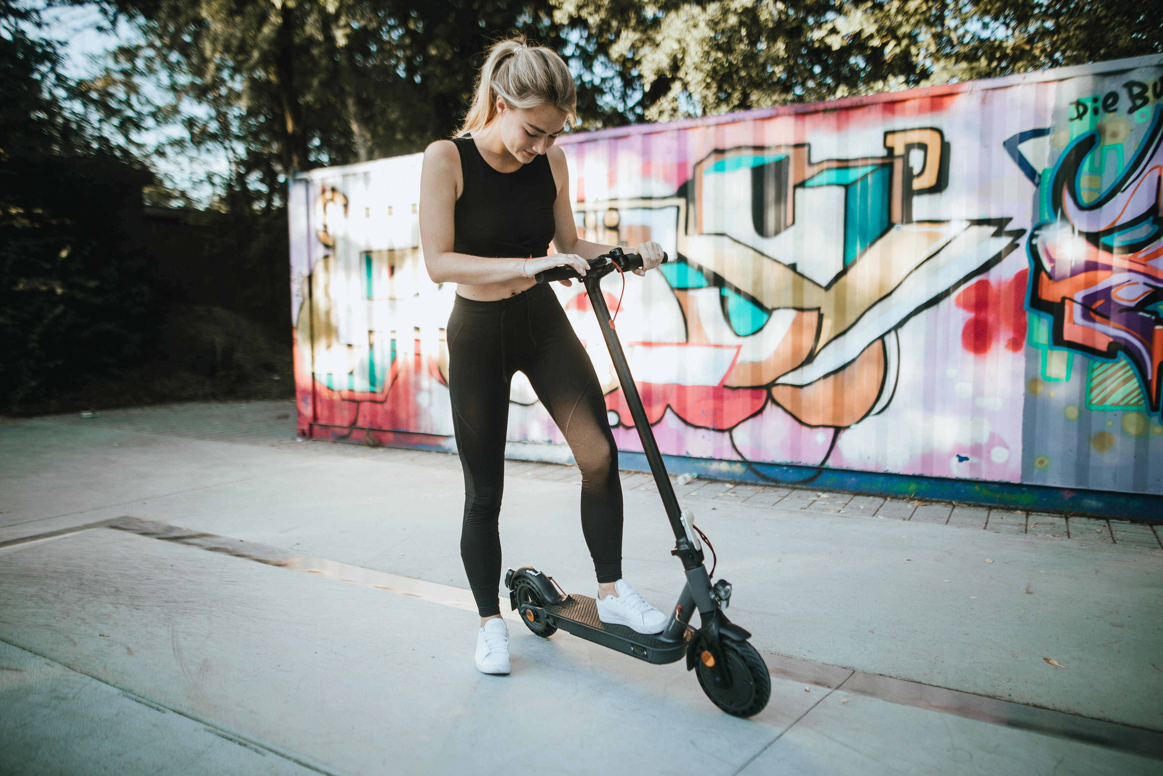 Junge Frau steht auf einem Roller auf einer Betonfläche, mit Graffiti im Hintergrund.