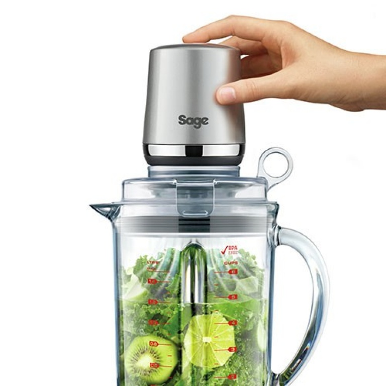 Una mano che posiziona un coperchio argentato su un estrattore di succo. Kiwi e lime verdi visibili.