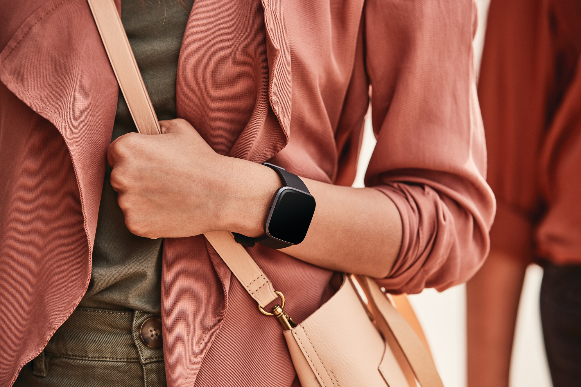Mujer con smartwatch, sosteniendo un bolso, de pie cerca de una chaqueta naranja.