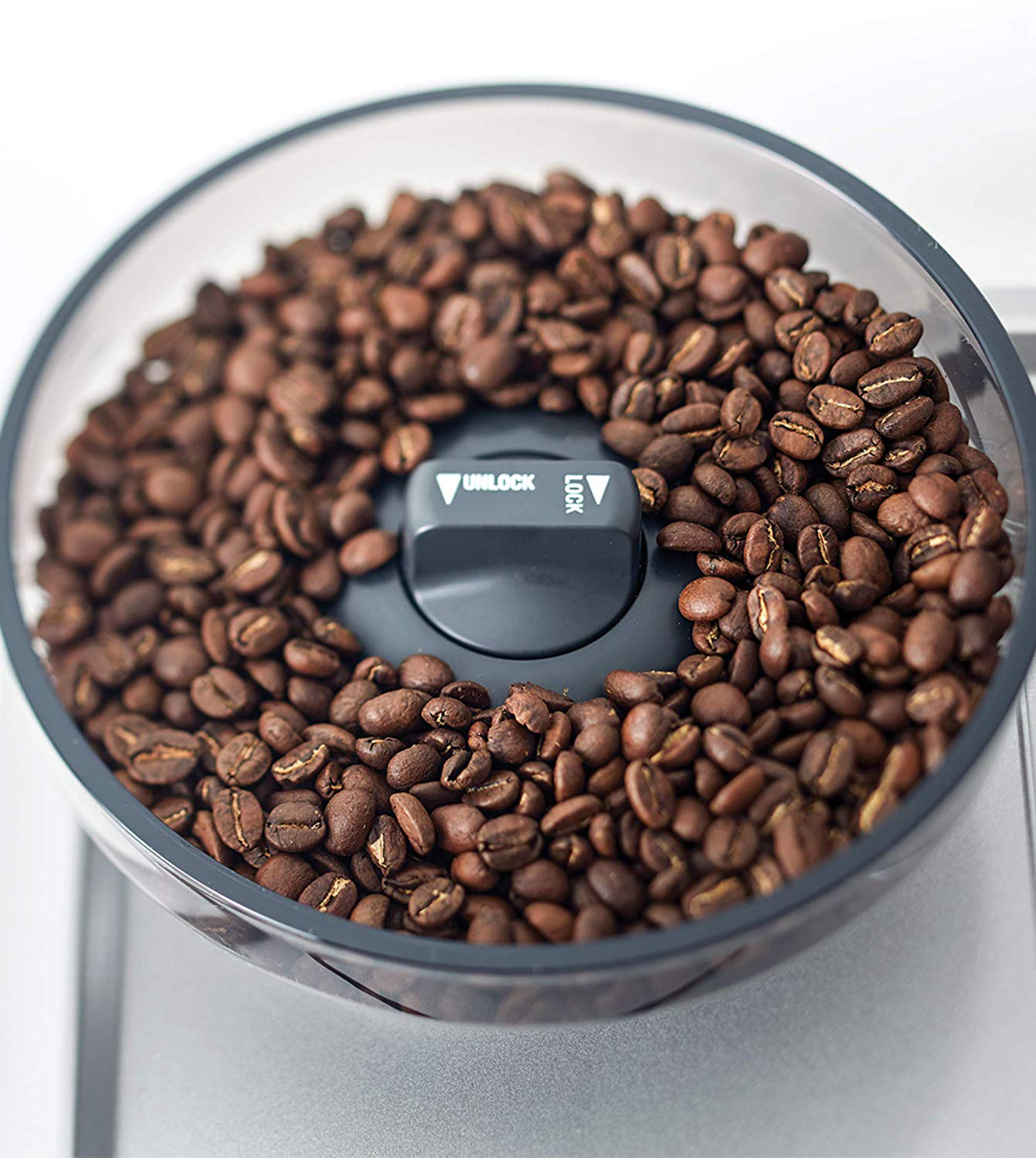 Un récipient transparent rempli de grains de café torréfiés. Un cadran noir avec des indicateurs de déverrouillage et de verrouillage.