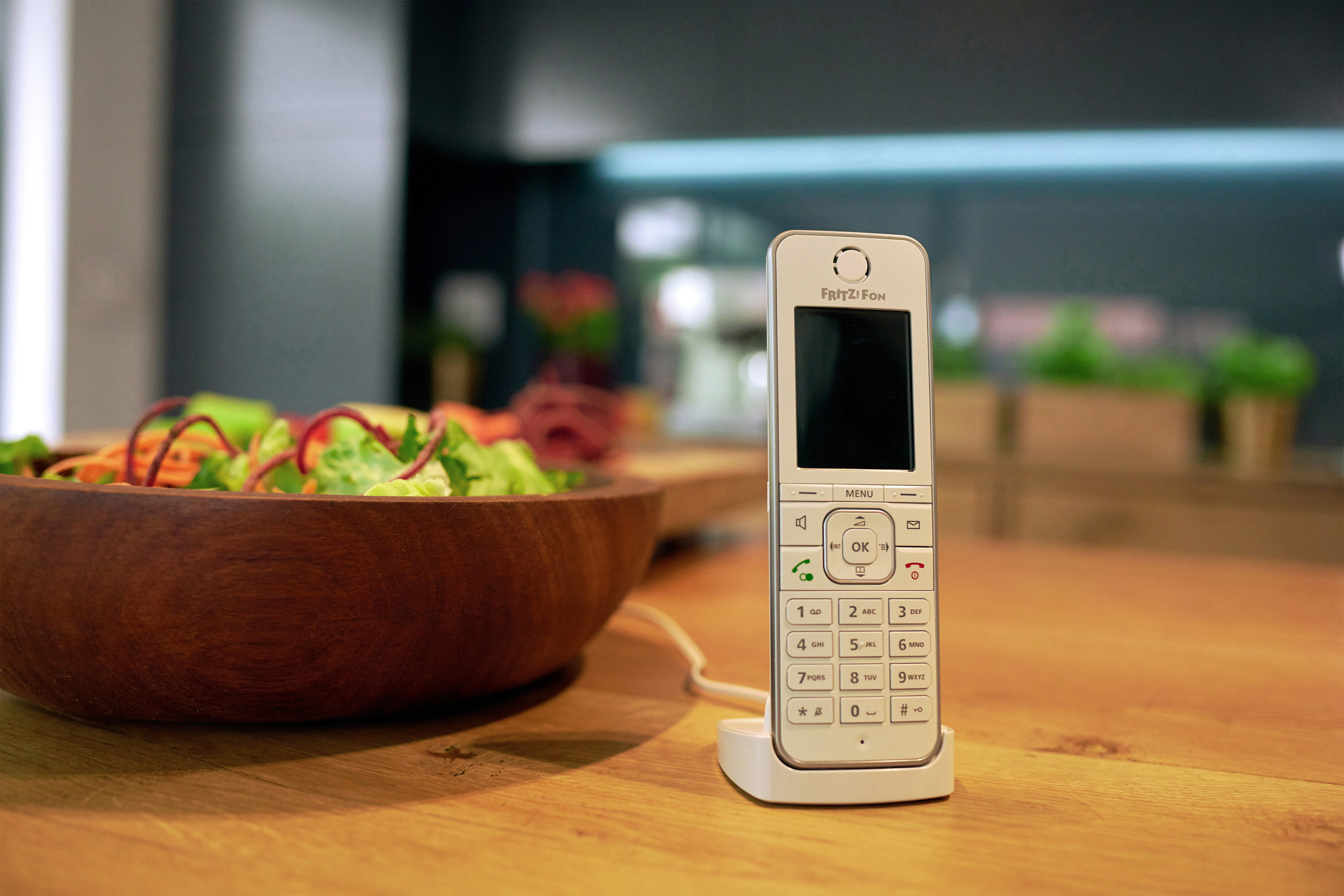 Weißes schnurloses Telefon auf einem Ladegerät. Ein Holzkrug mit Salat ist im Hintergrund.