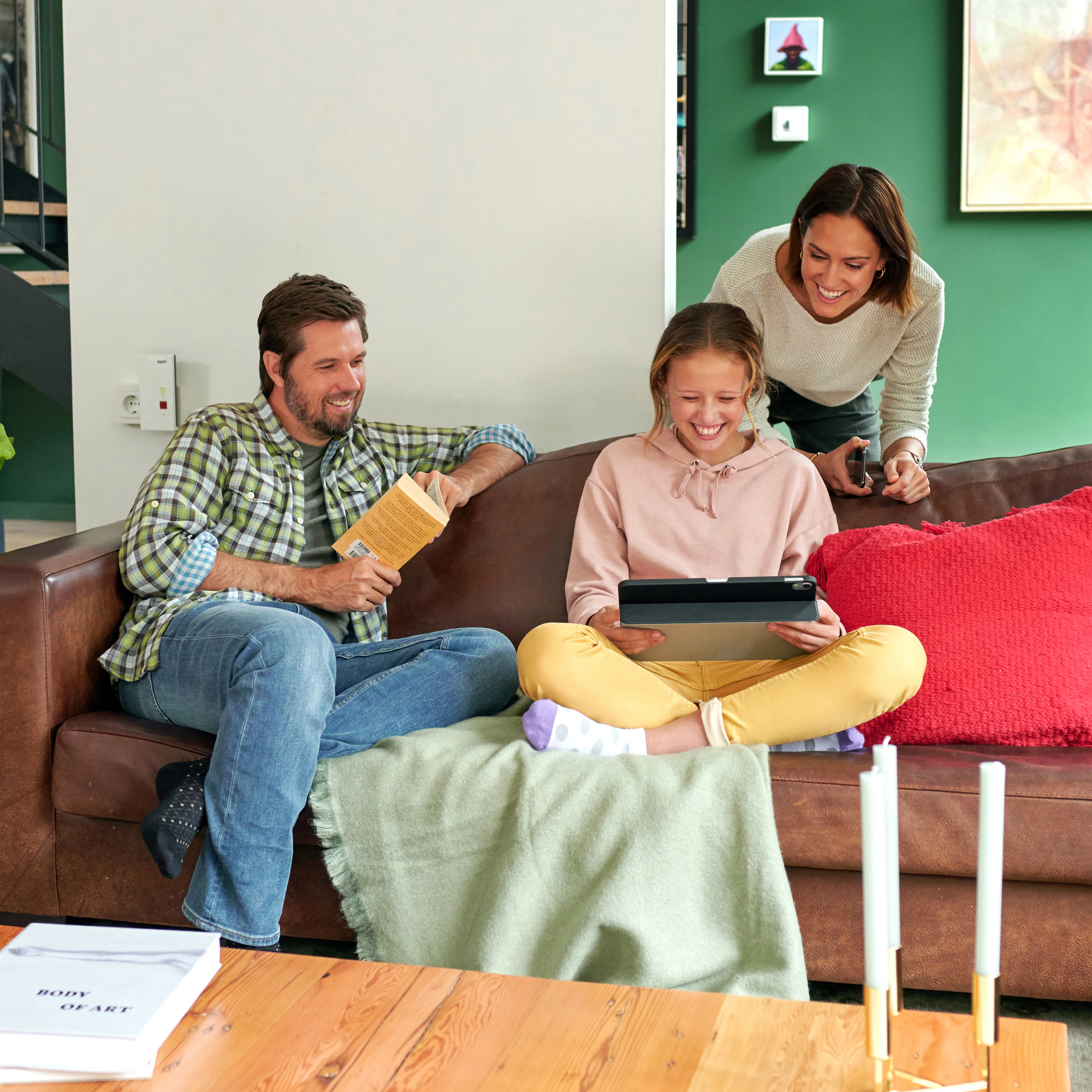 Familie auf einem braunen Sofa. Vater liest, Tochter mit Tablet, Mutter steht. Grüne und rote Akzente.