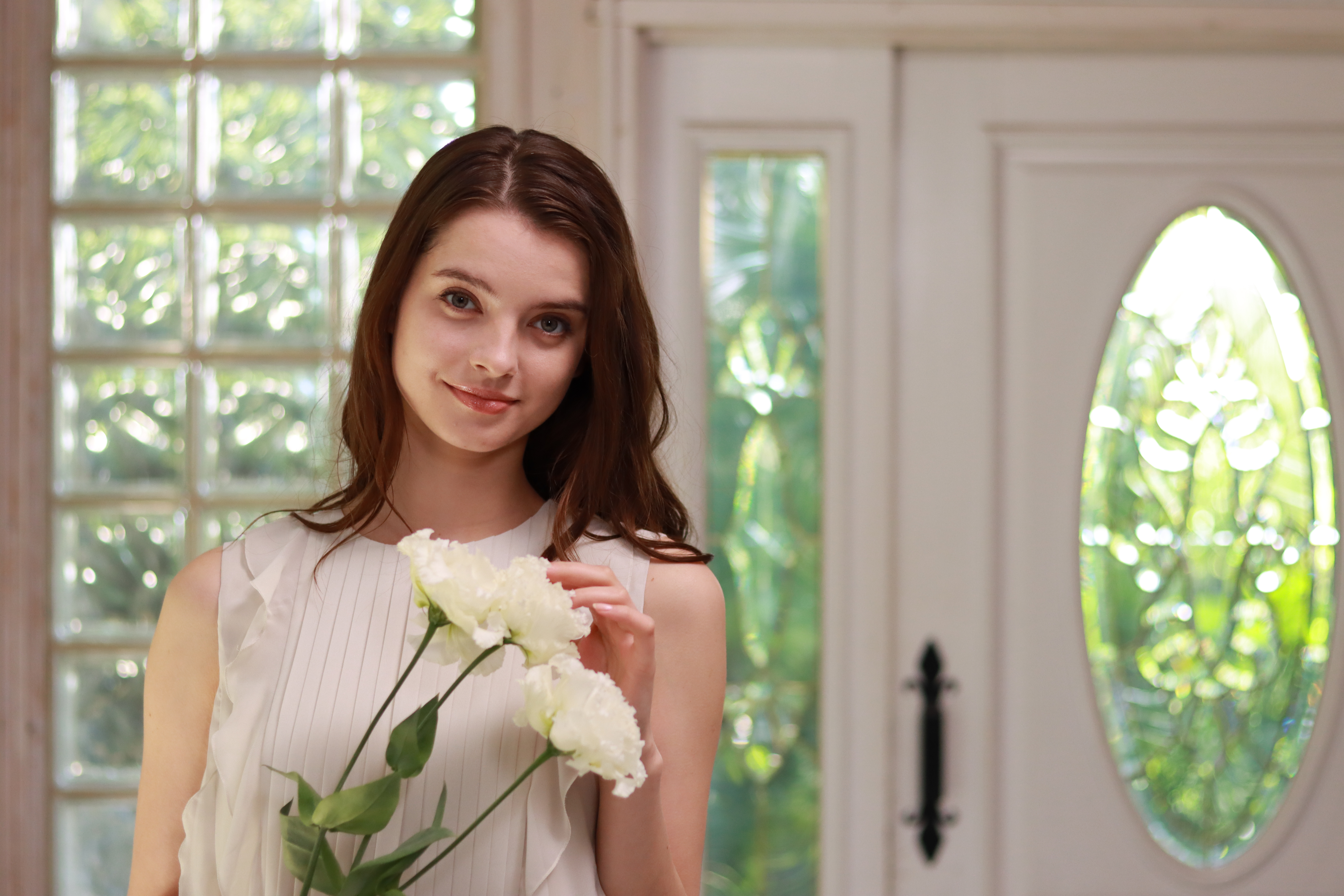 Eine Frau hält weiße Blumen und lächelt in die Kamera. Der Hintergrund ist eine Glastür mit weißem Rahmen.