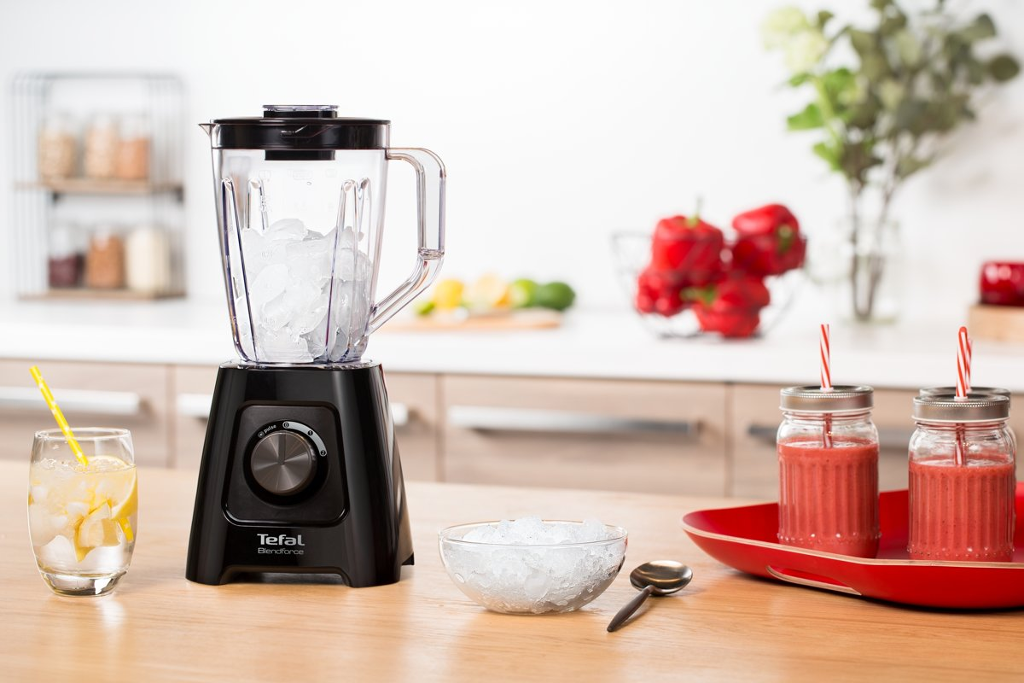 Egy fekete turmixgép jéggel, hozzávalókkal és piros smoothie-kkal, egy fa asztalon.