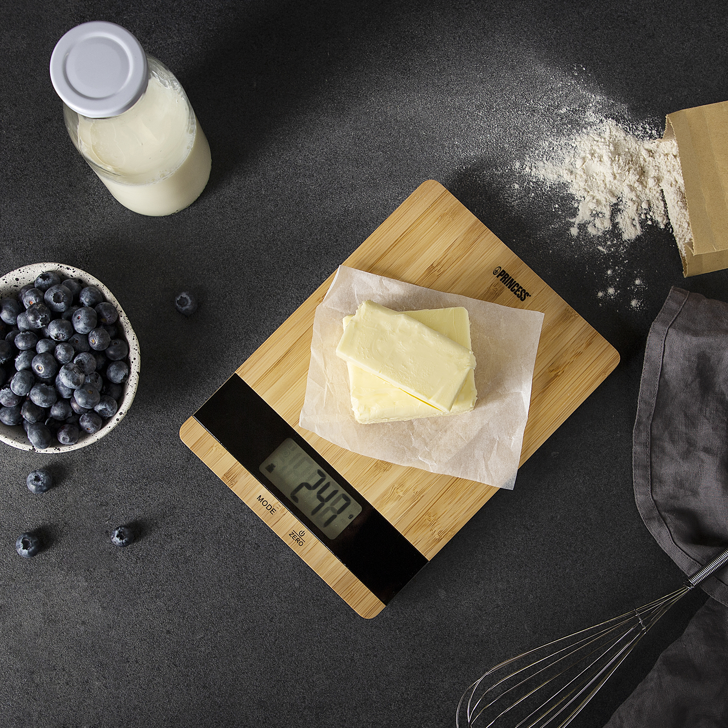 Ingredients on a black surface: blueberries, milk bottle, butter, flour, and a scale. 