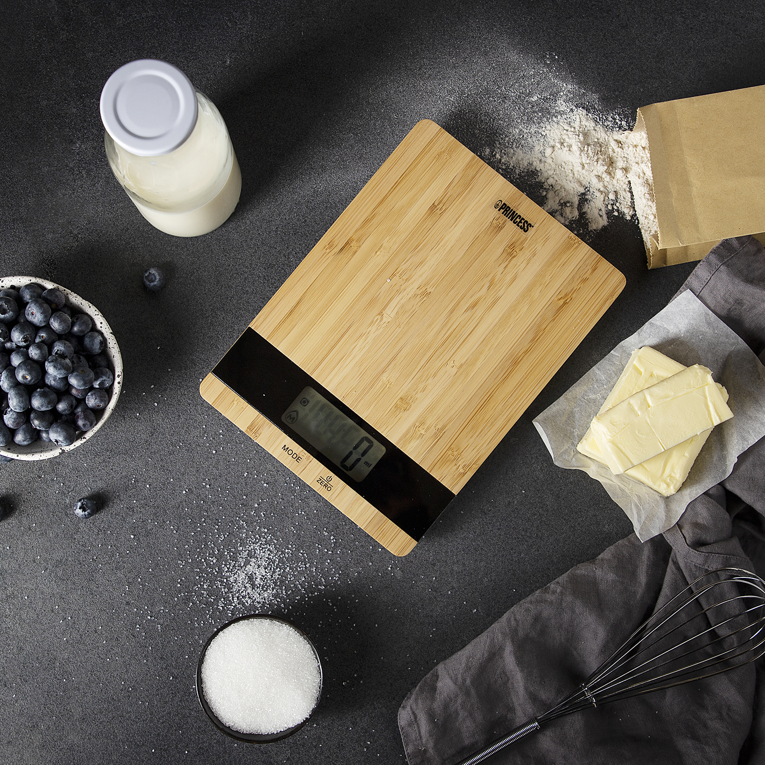 A bamboo food scale on a dark surface, with ingredients like blueberries, sugar, and butter around it.