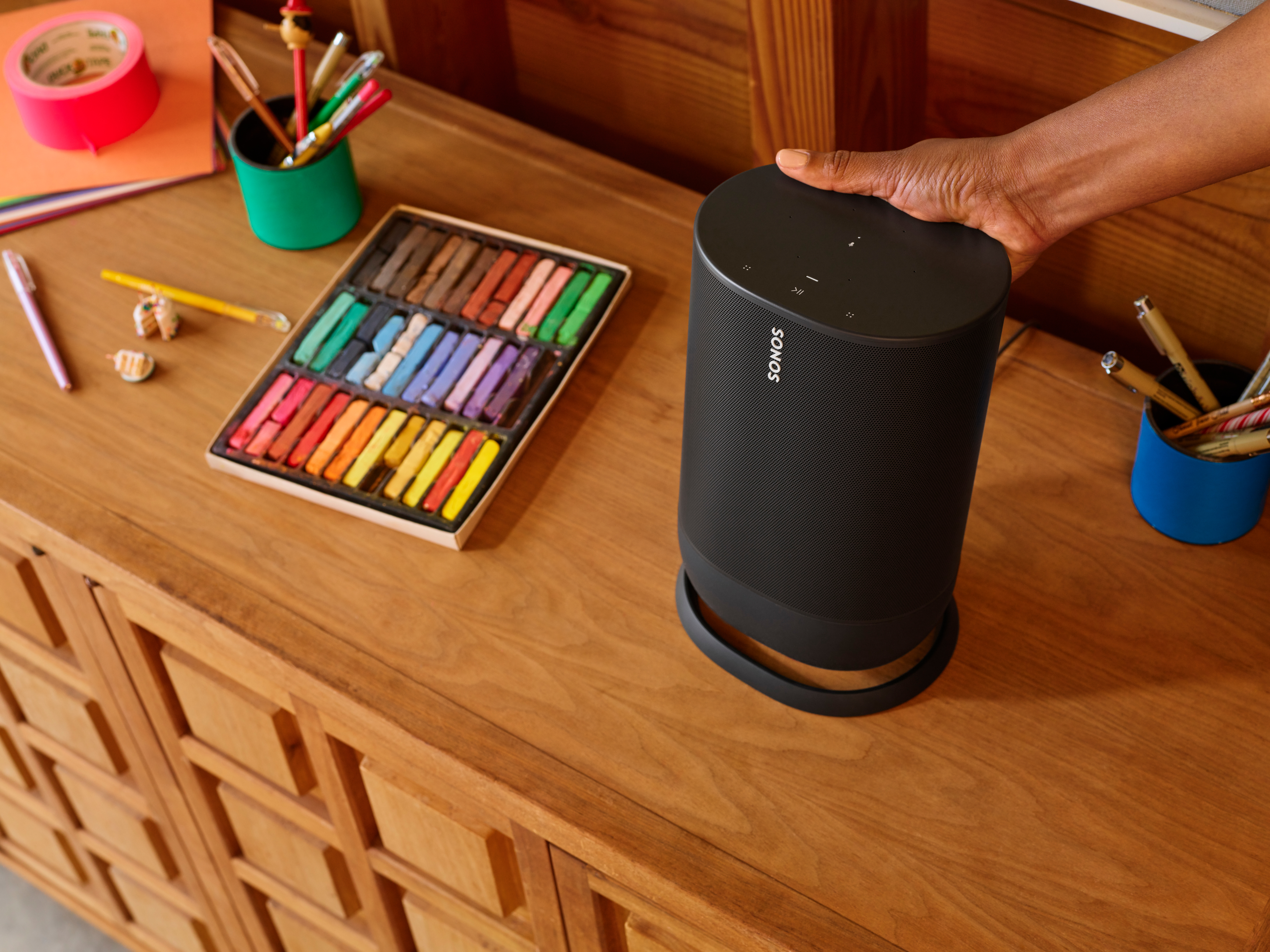 Ein schwarzer Sonos-Lautsprecher, der von einer Hand berührt wird. Verschiedene Künstlerutensilien auf einem Holzschrank.
