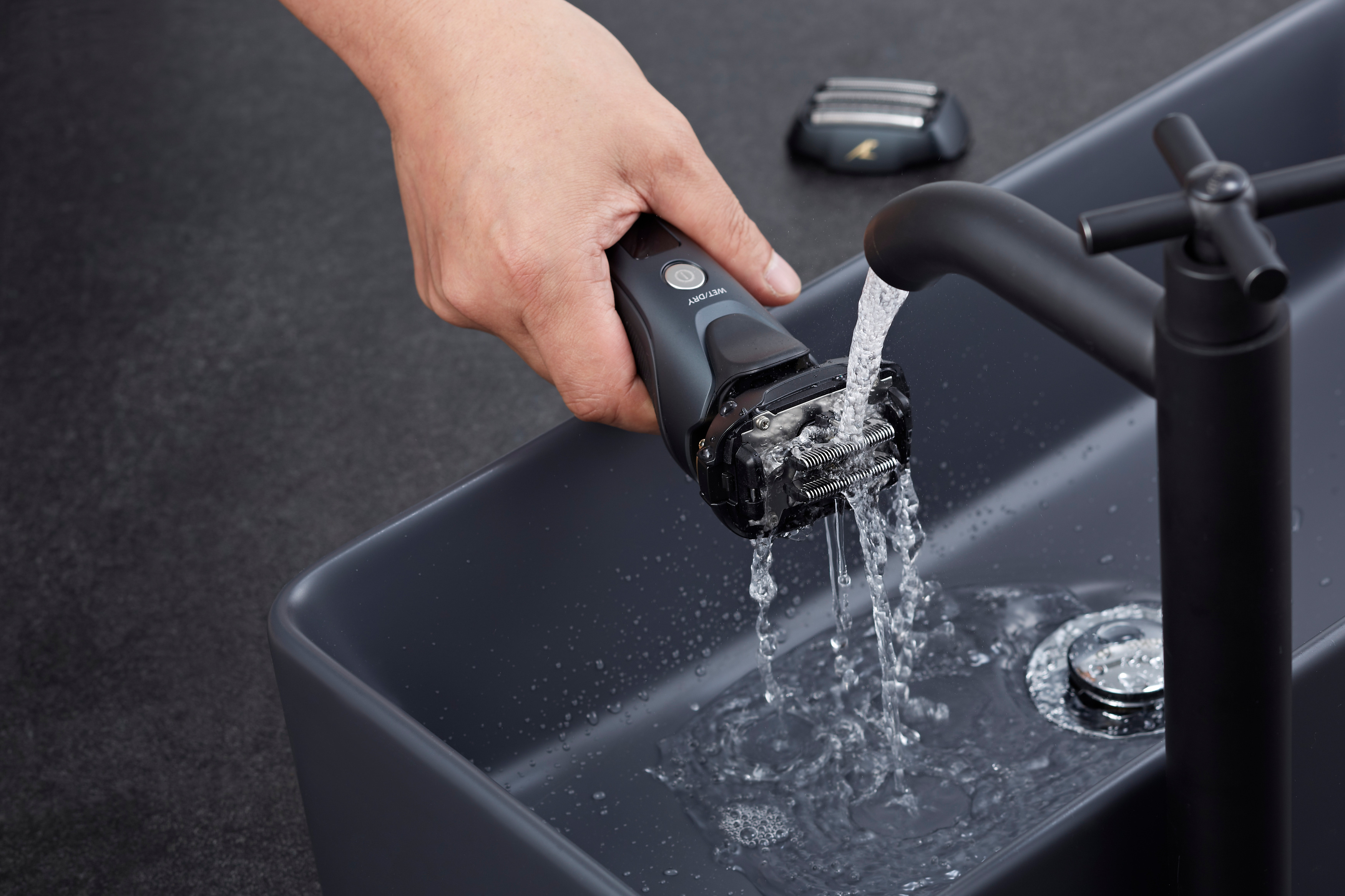 Eine Hand hält einen schwarzen Elektrorasierer unter fließendem Wasser in einem grauen Waschbecken.