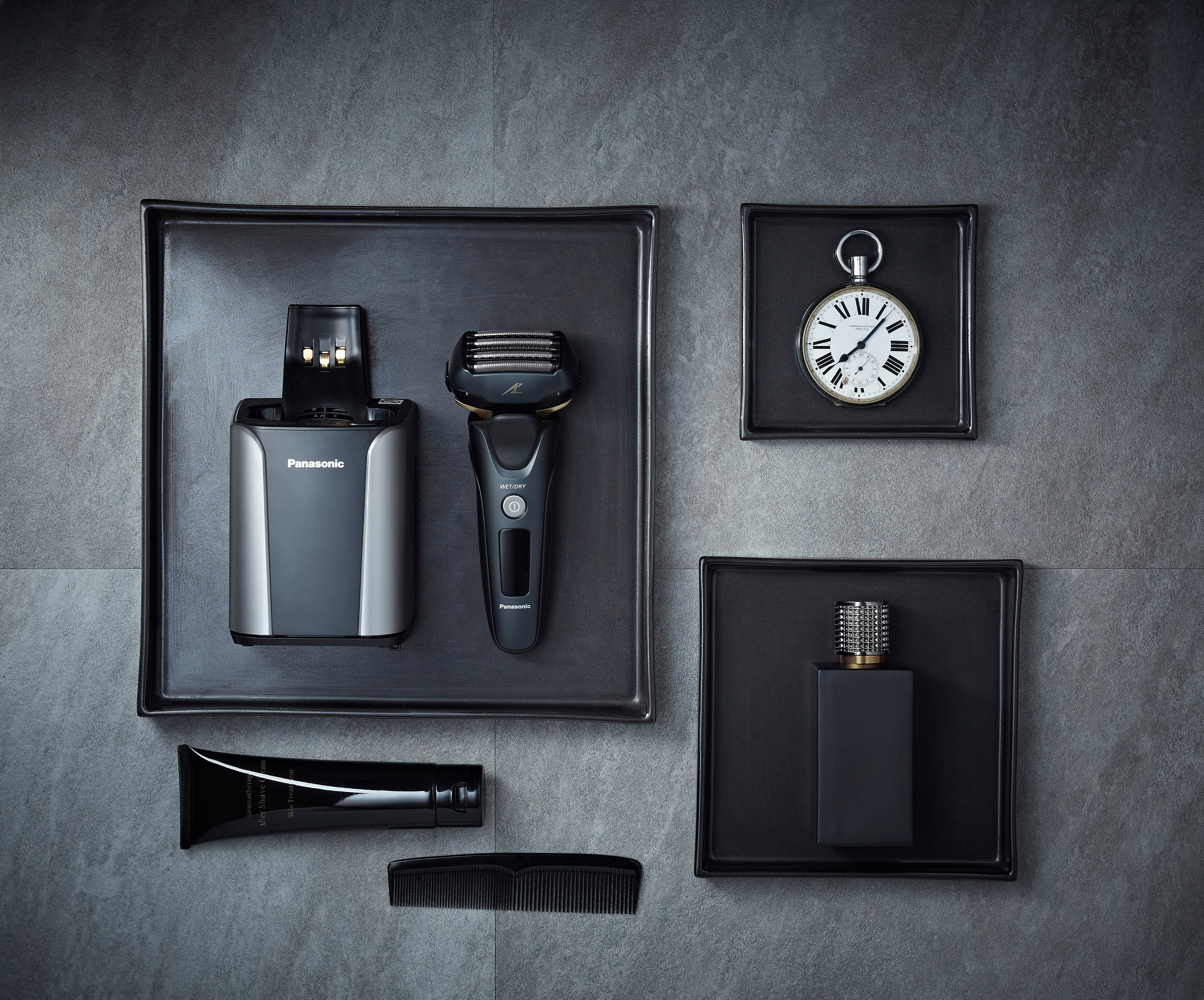 Verschiedene Pflegeutensilien auf schwarzen Tabletts: Rasierer, Creme, Kamm und Uhr.