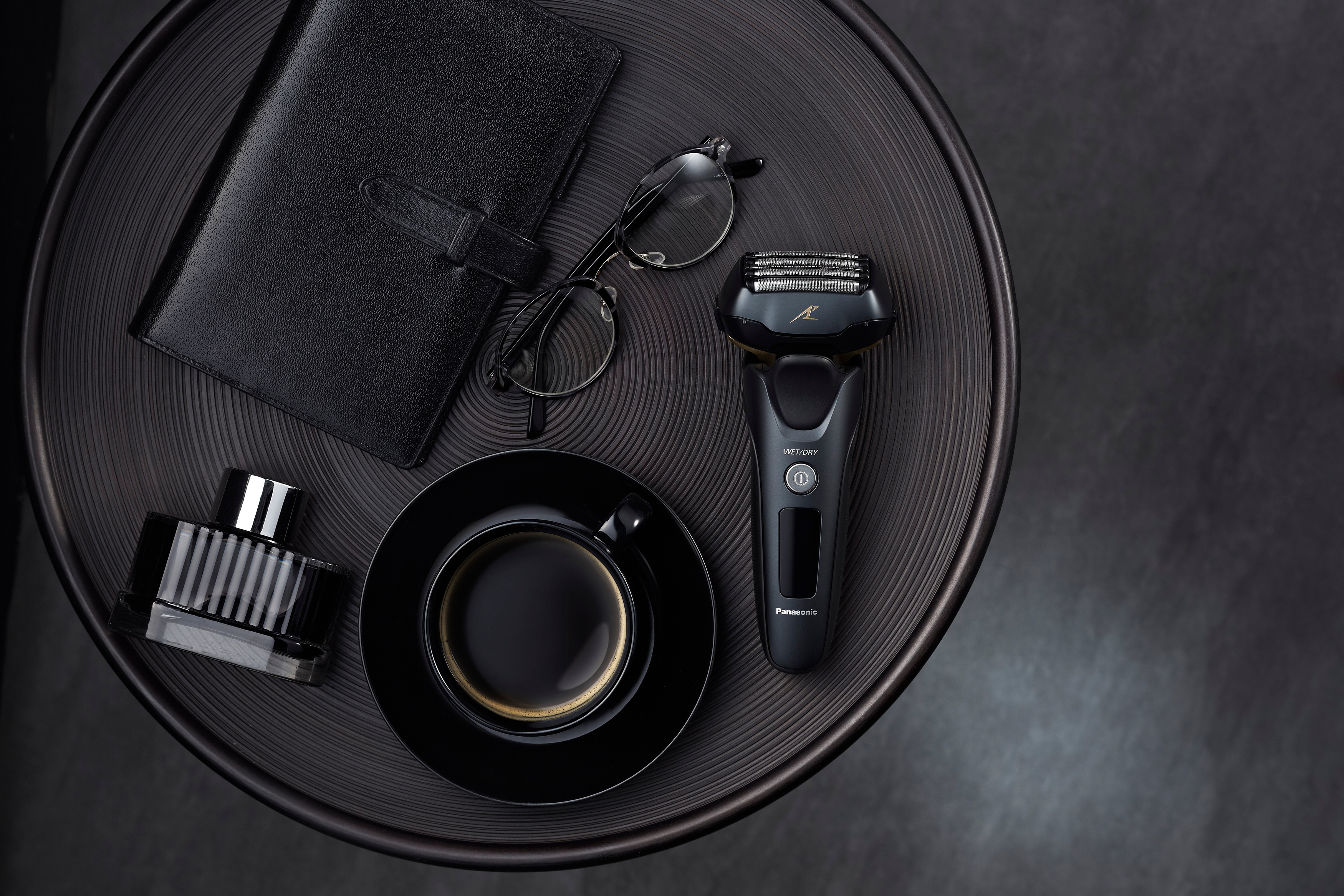 Schwarzes Pflege-Set auf einem dunklen Tisch mit Notizbuch, Brille, Parfüm und Kaffee.