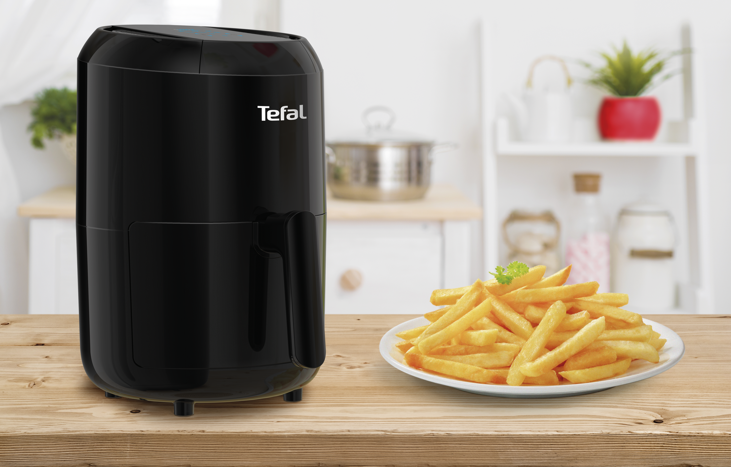 A black Tefal air fryer on a wooden table with fries on a plate and kitchen in the background.