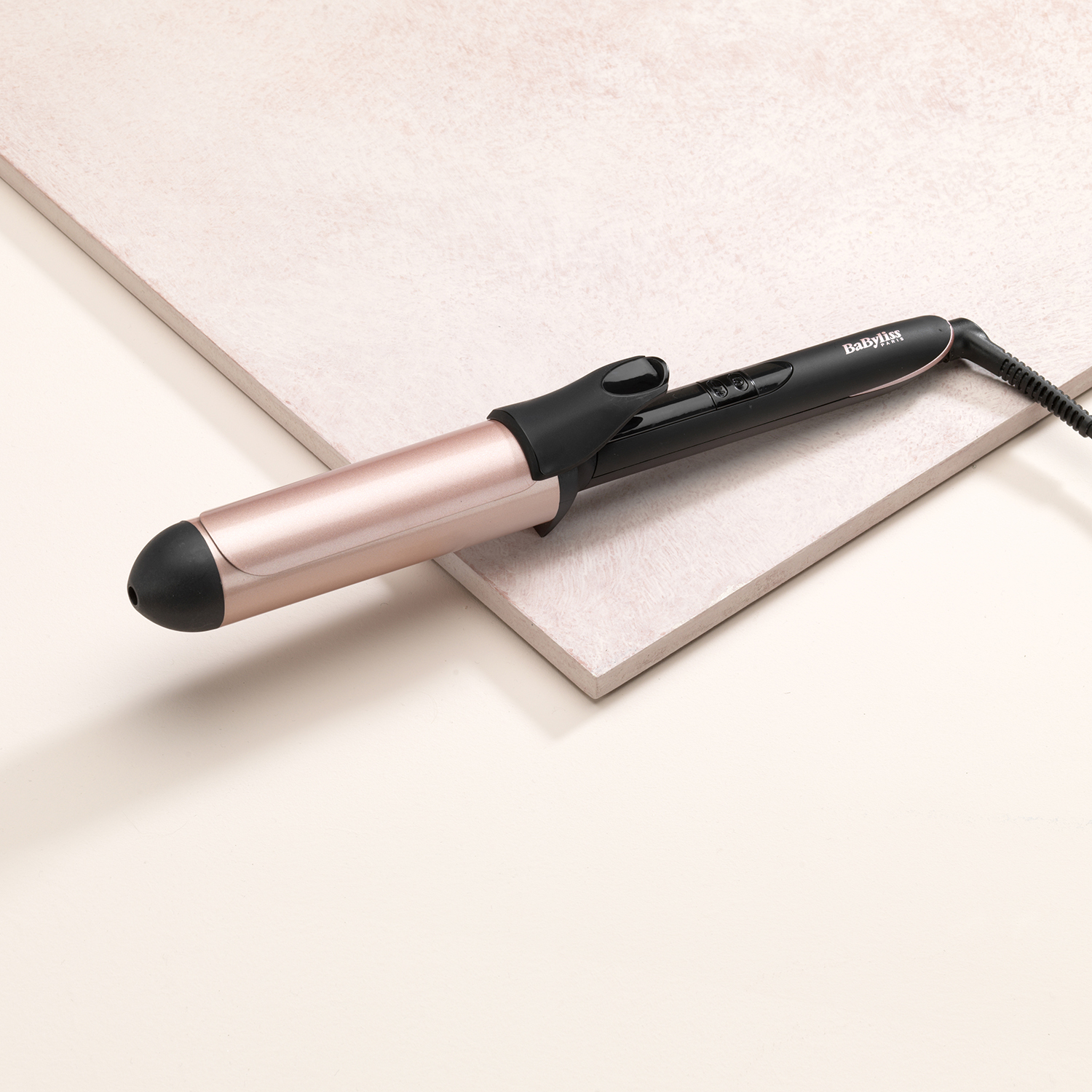 A curling iron with a black handle and a rose gold barrel, sitting on a pink surface.
