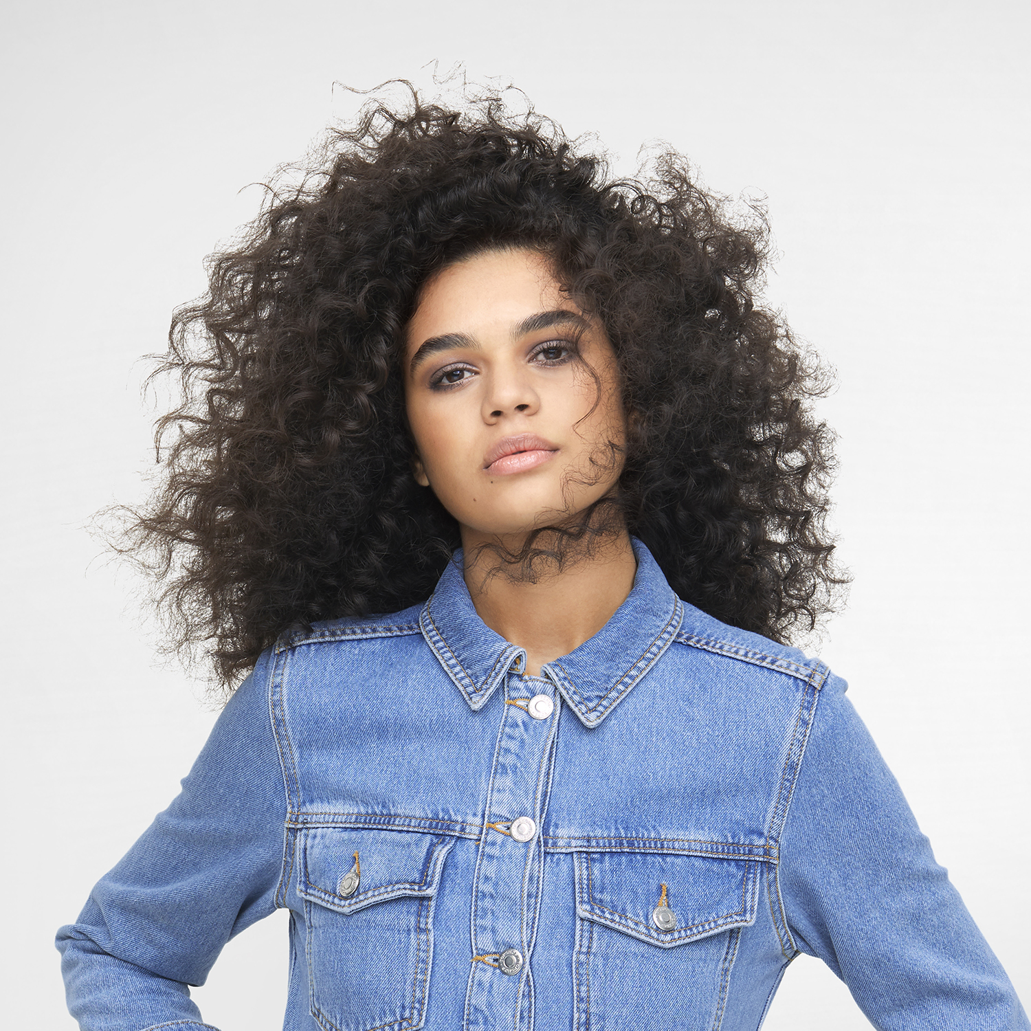 Woman with curly hair, wearing a denim jacket, looking at the camera.