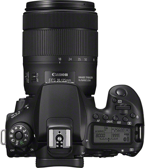 Top-down view of a Canon DSLR camera. Black body, lens attached, many buttons, white background.