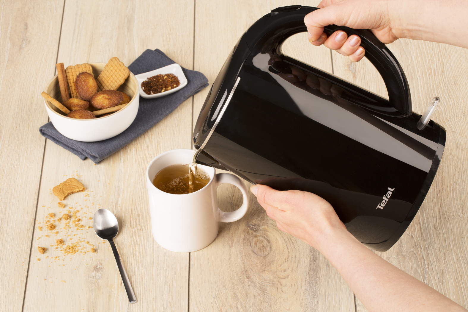 A person pouring tea from a black kettle into a white mug. Biscuits and spices are in the scene.
