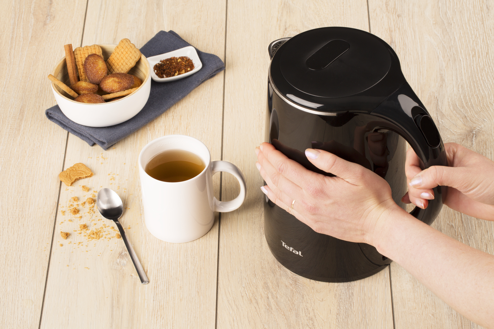 Woman holding a black kettle with a cup of tea and biscuits on a table.